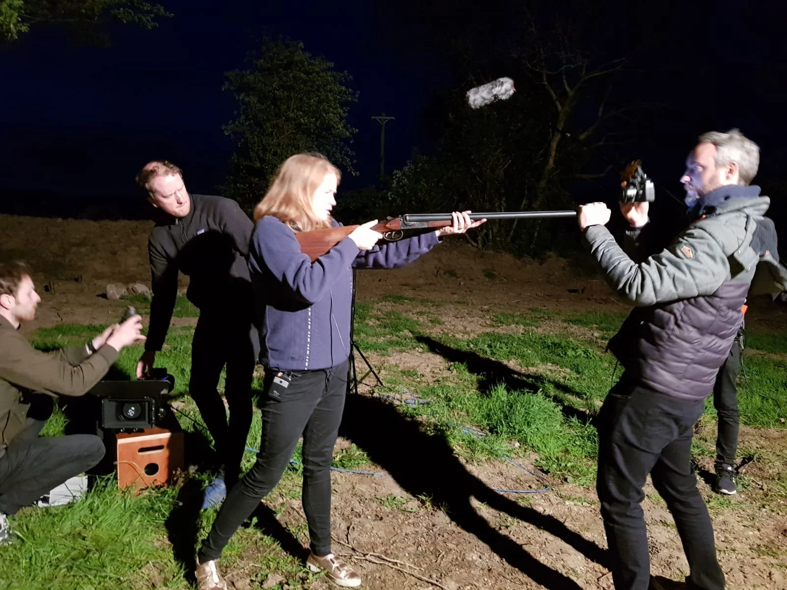 A behind the scenes photo. A woman aiming a rifle at a shooting target, with a man holding a camera on her right.