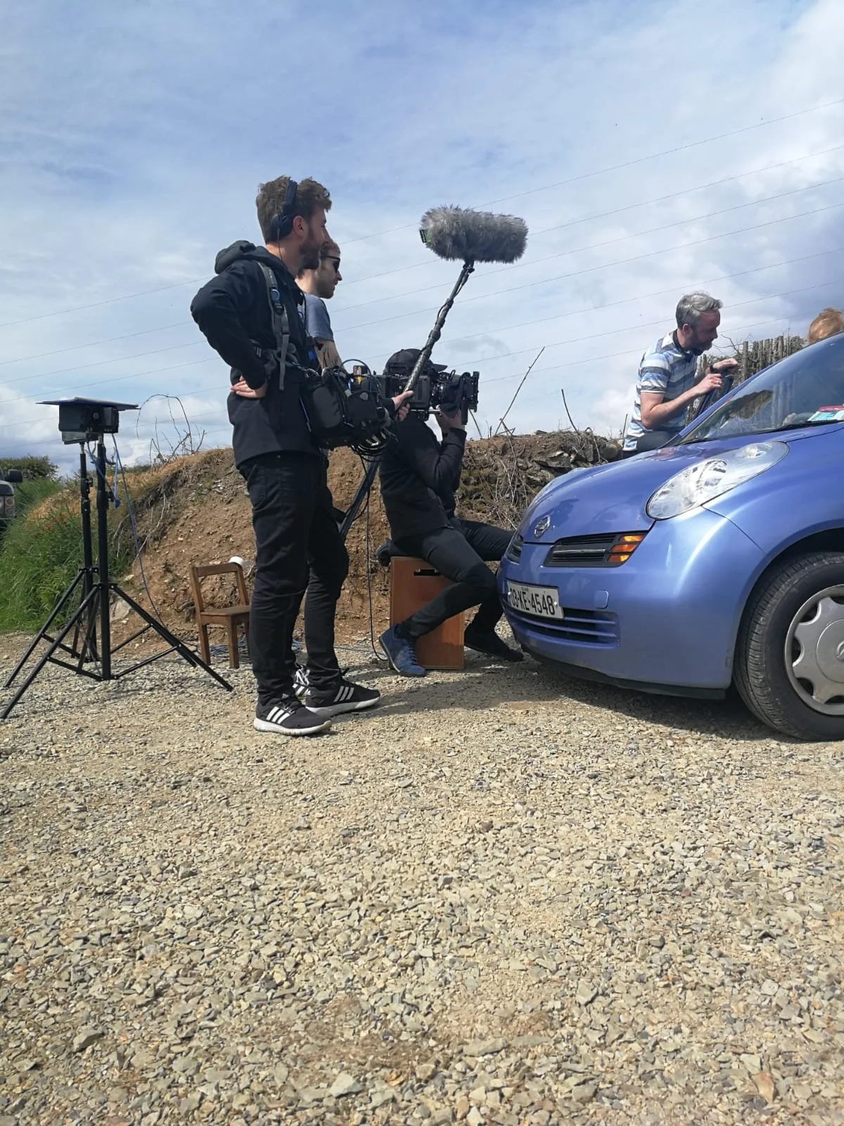 A film crew using a camera and audio equipment film a scene with a man sitting behind a blue car, with another man adjusting something near the car's windshield, on an outdoor gravel lot.