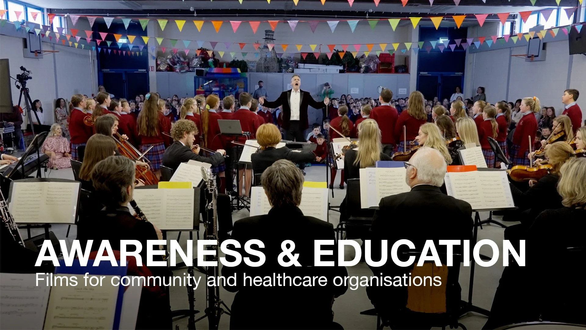 A large indoor concert with a conductor leading an orchestra of students, with a choir of students in red uniforms standing behind them. The audience is watching while colorful flags hang from the ceiling. The scene appears lively and organized for a