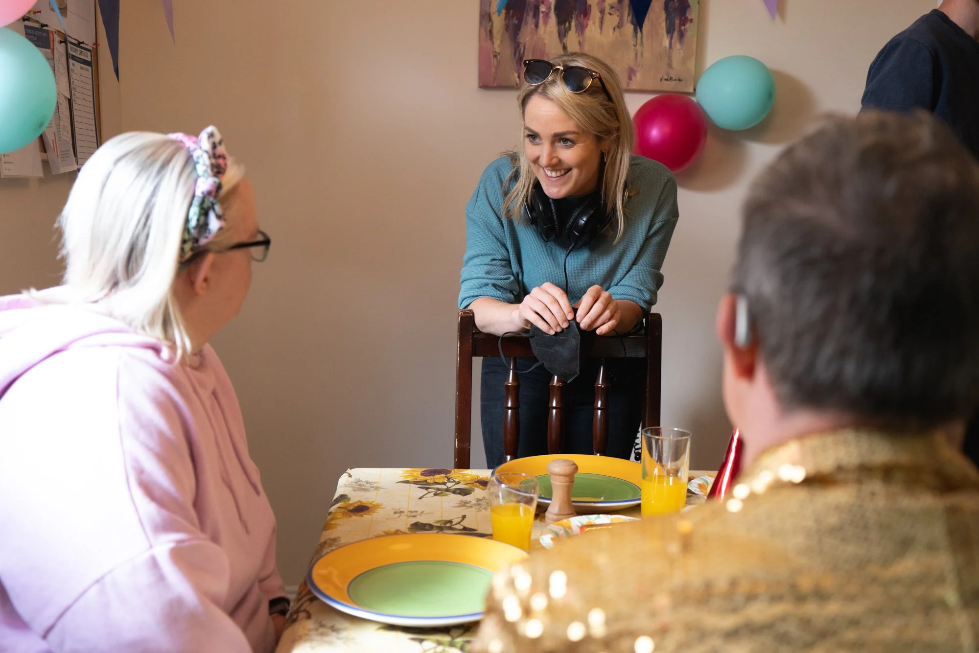 Behind the scenes photo. Smiling woman with blonde hair, sunglasses on her head, and headphones around her neck, standing behind a dining table with party balloons on the wall, talking to a group of people at a celebration.