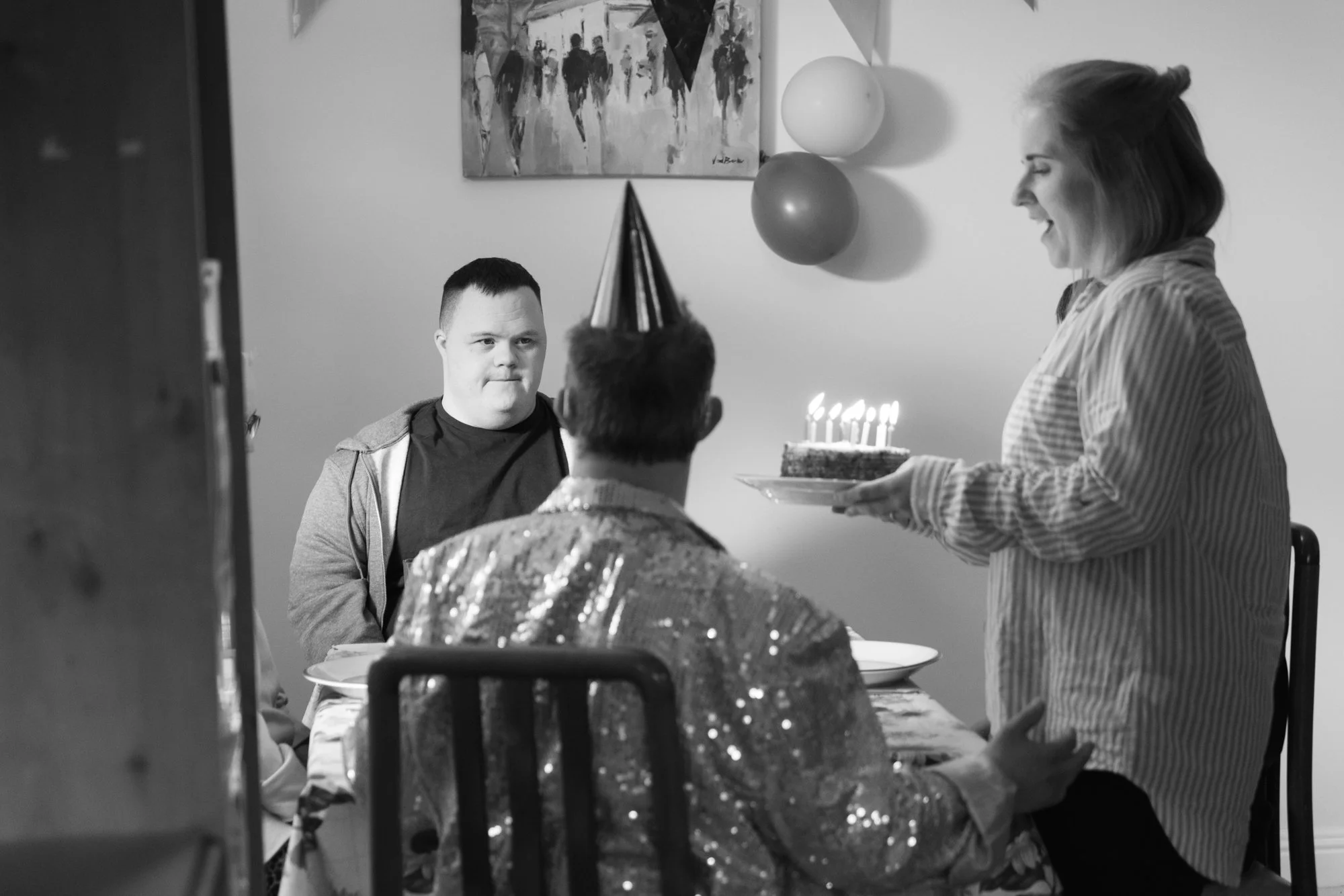 Behind the scenes photo from a film set. Woman smiling and holding birthday cake with lit candles at a table where people are gathered for a celebration.