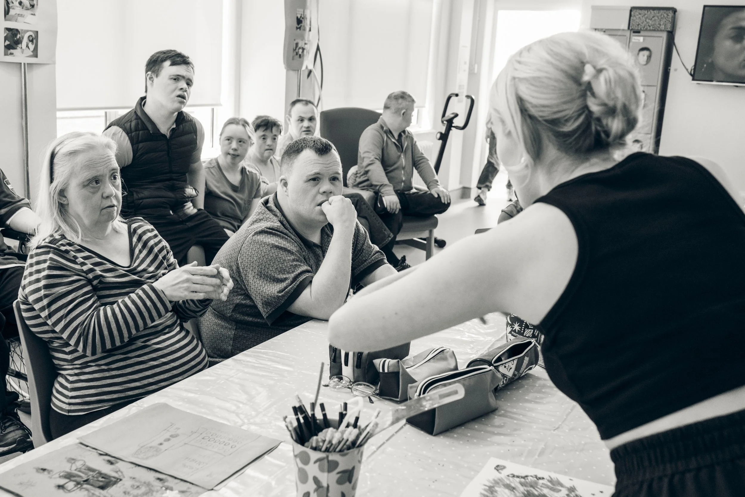A group of children and adults sitting around a table in a classroom or therapy room, listening to a woman standing at the front, engaging in conversation.