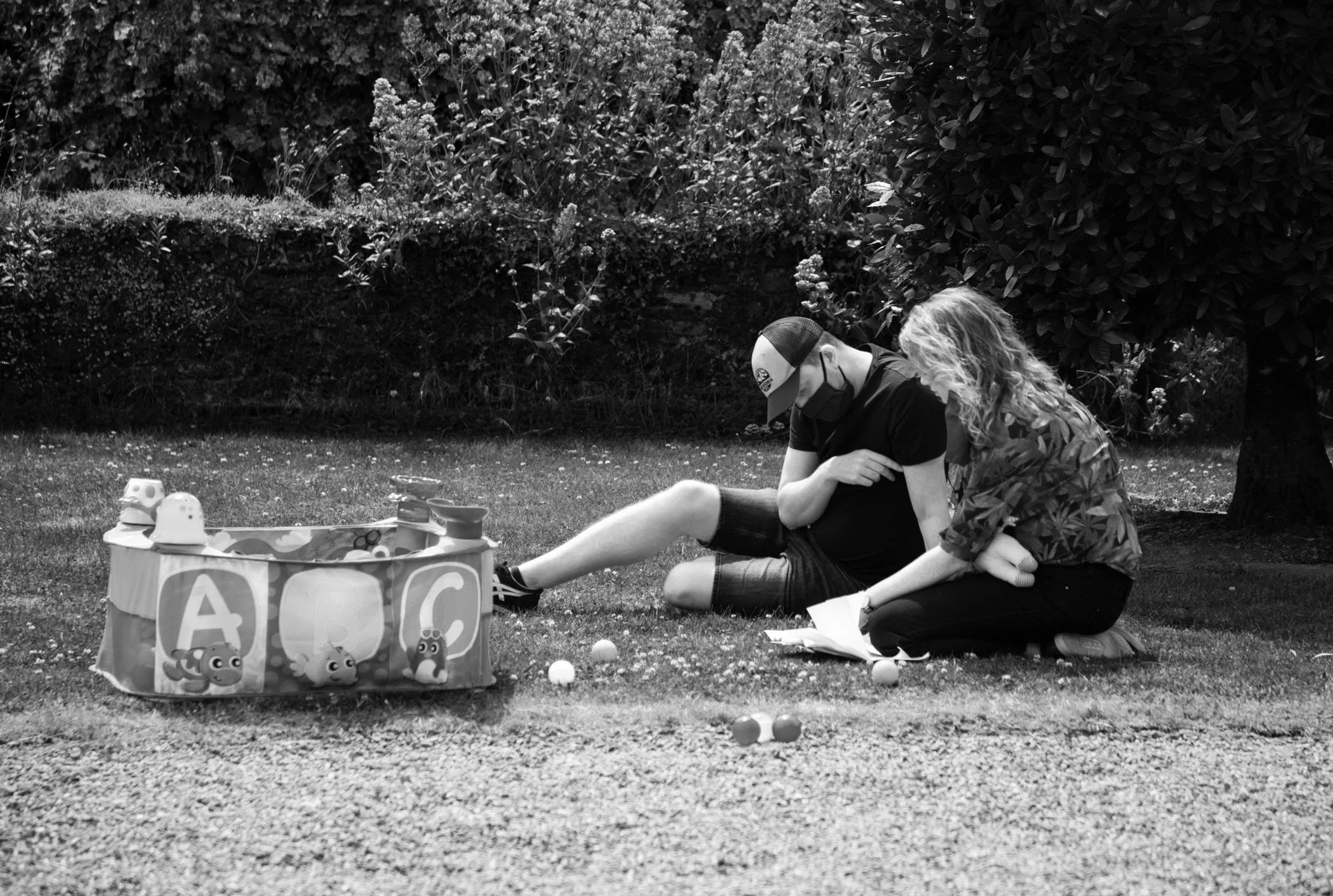 Behind the scenes photo. Two children sitting on the grass, playing a game with plastic balls. There is a toy bucket with animal stickers and some balls nearby. They are outdoors with shrubs and trees in the background.