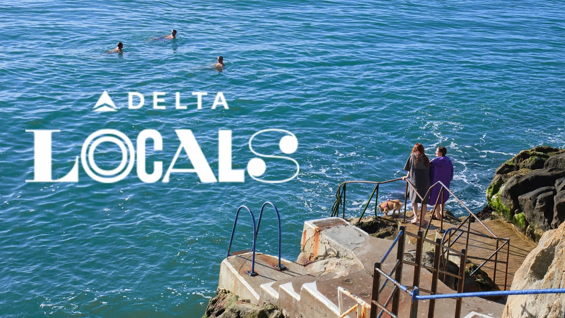 Two women with a dog standing on a rocky pier looking out at the ocean, with swimmers in the water and a 'Delta Local' overlay.