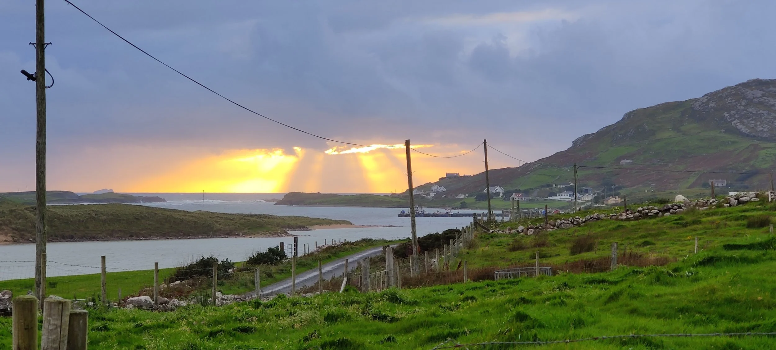 Scenic view of a bay with a small island, with sunlight rays breaking through clouds at sunset, green hills, houses, and a mountain in the background.