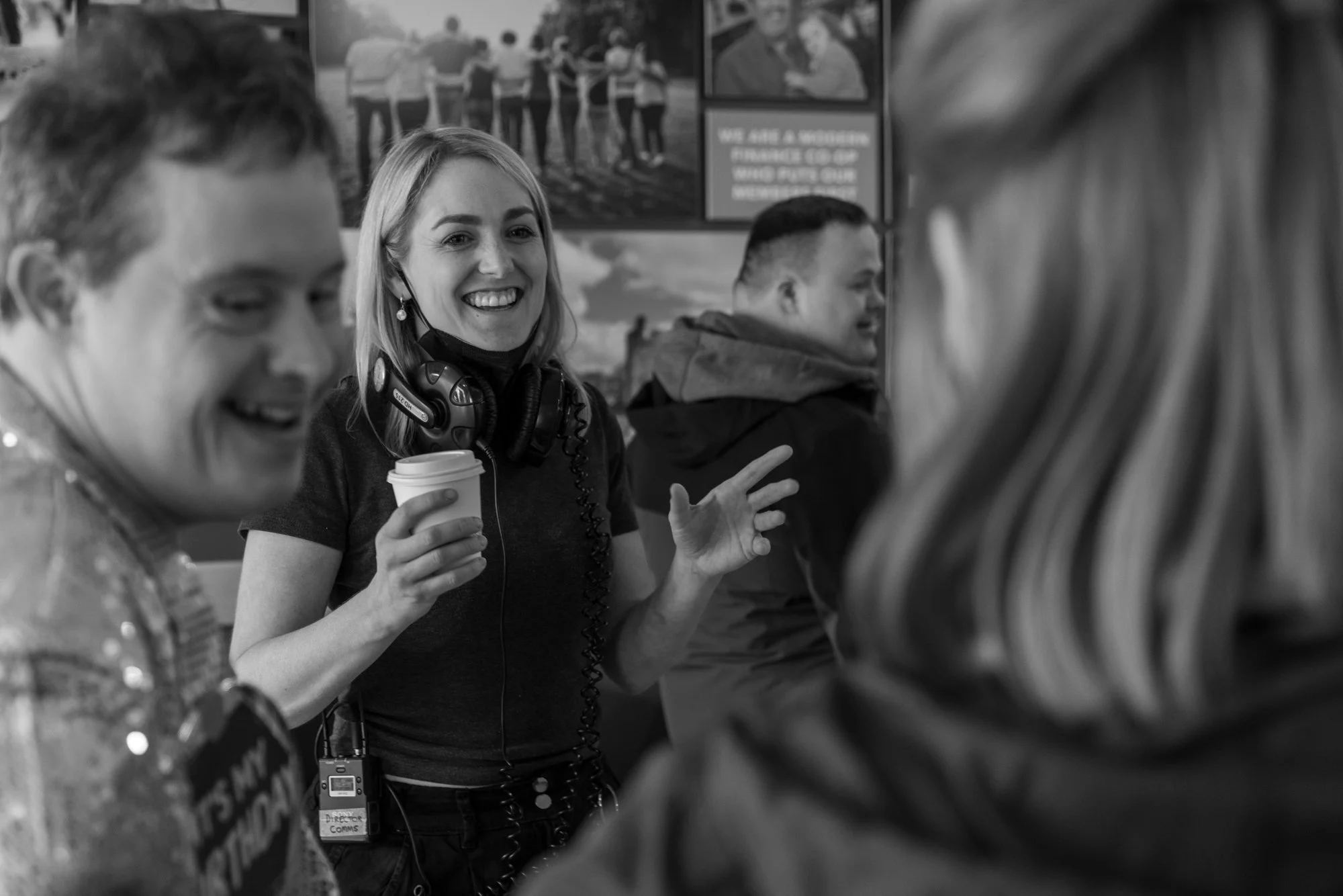 Behind the scenes photo. A woman with blonde hair smiling and holding a cup of coffee, wearing a headset, standing among a group of people in a busy indoor setting with posters on the wall.