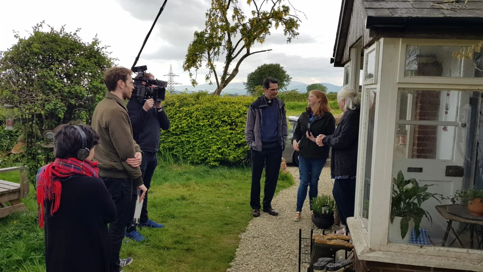 Filming crew and cast members are filming a scene outdoors next to a small house, with a woman in black being interviewed by another woman, amid overcast weather.