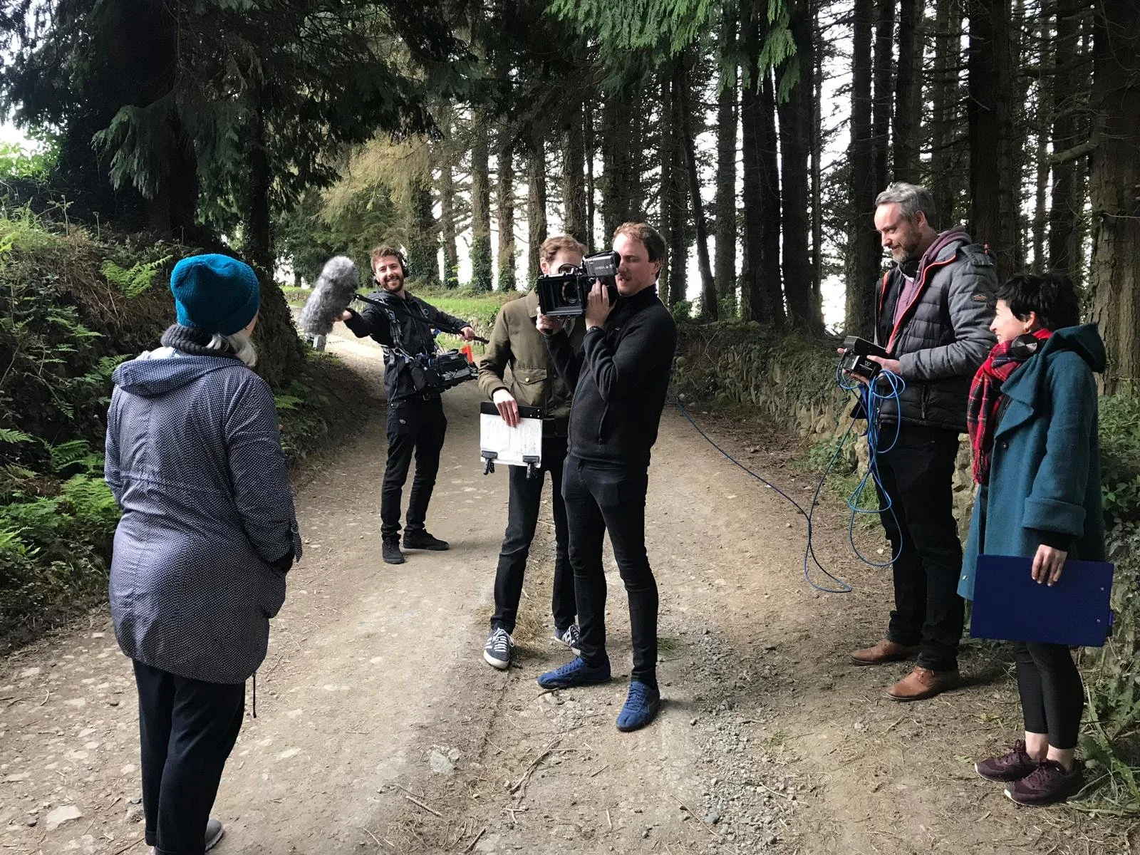 Film crew filming in a forest with trees and a dirt path, including a person speaking to the camera and crew members with equipment.