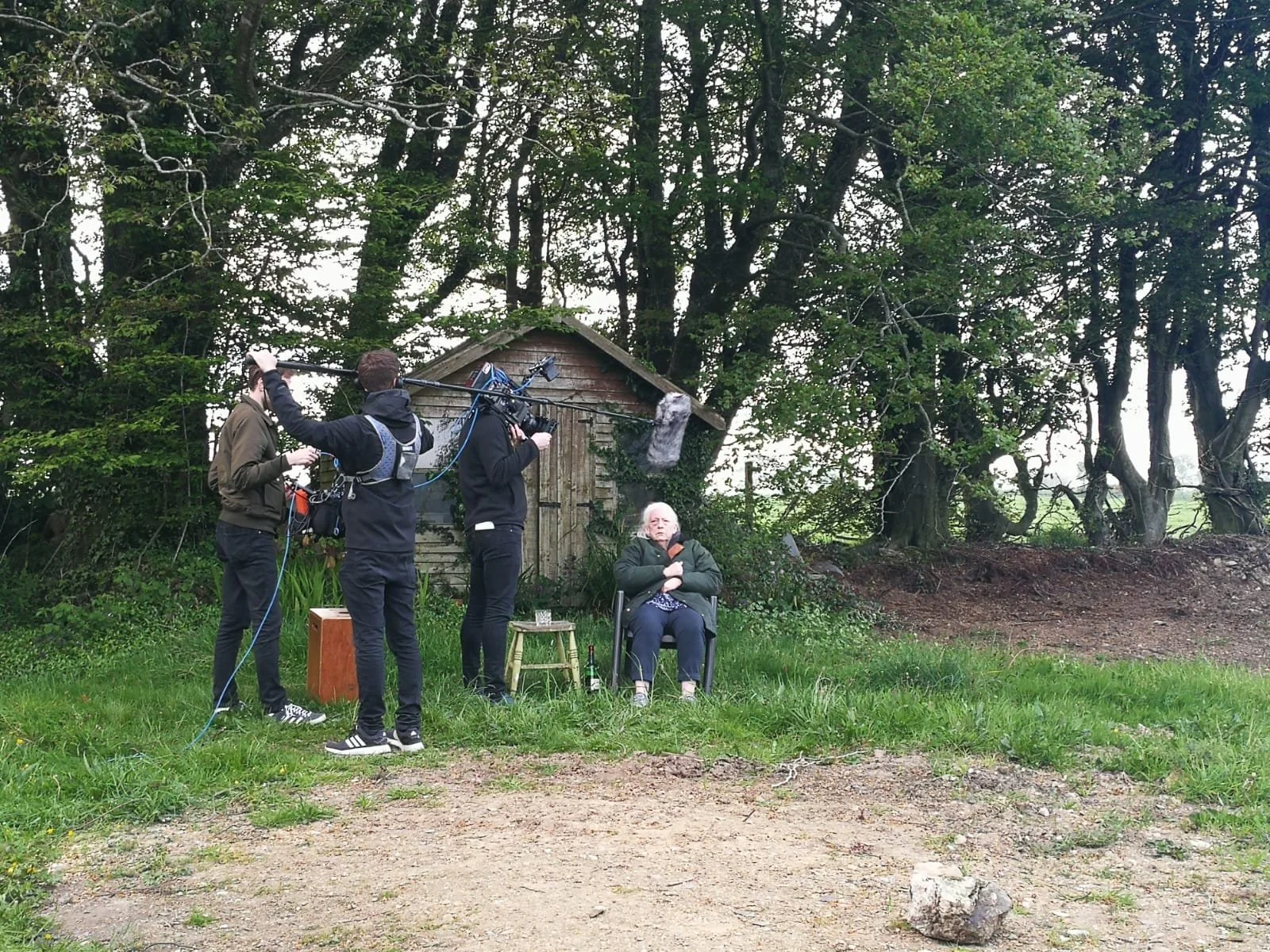 Behind the scenes of a video or film shoot in a lush, green outdoor setting with large trees, an old shed, and an elderly woman sitting in a chair, surrounded by film crew members operating equipment.