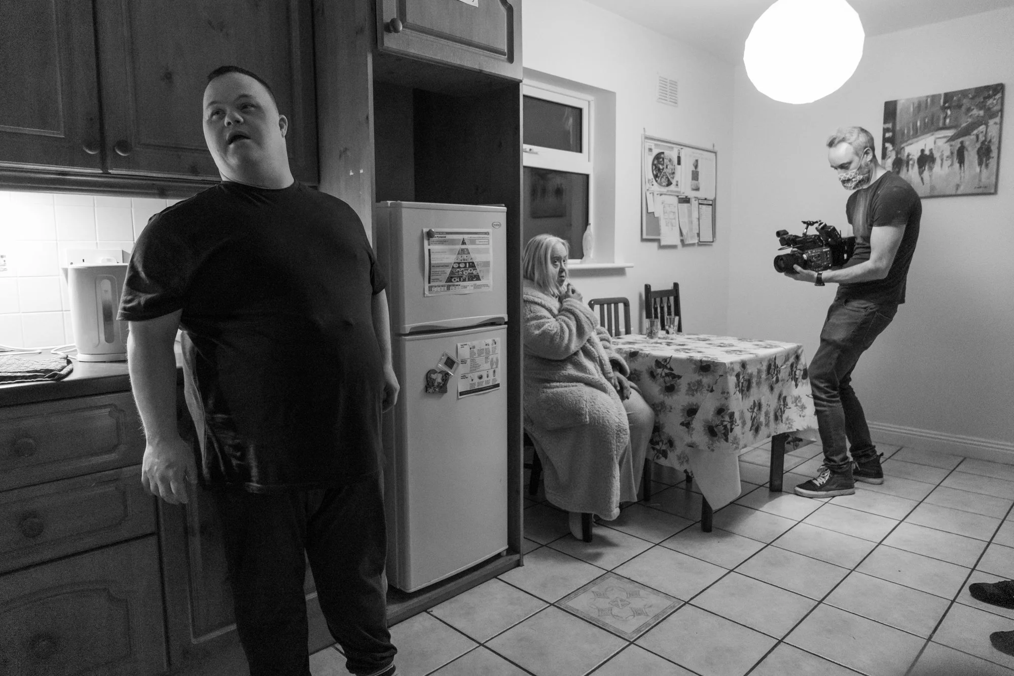 A woman sitting at a dining table with a floral tablecloth, wearing a hoodie and looking to her left, while a man stands nearby in the kitchen. Another man wearing a face mask and glasses is holding a professional video camera on the right side of th
