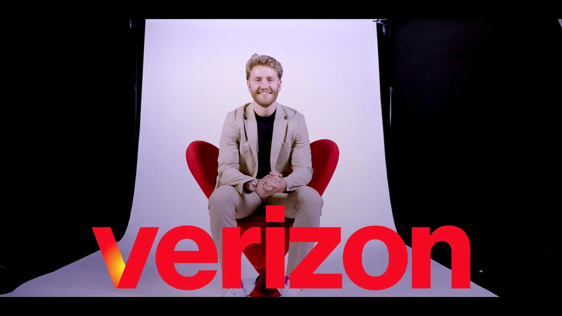 A smiling man sitting on a red chair in front of a white backdrop with the word 'Verizon' in large red letters in front of him.
