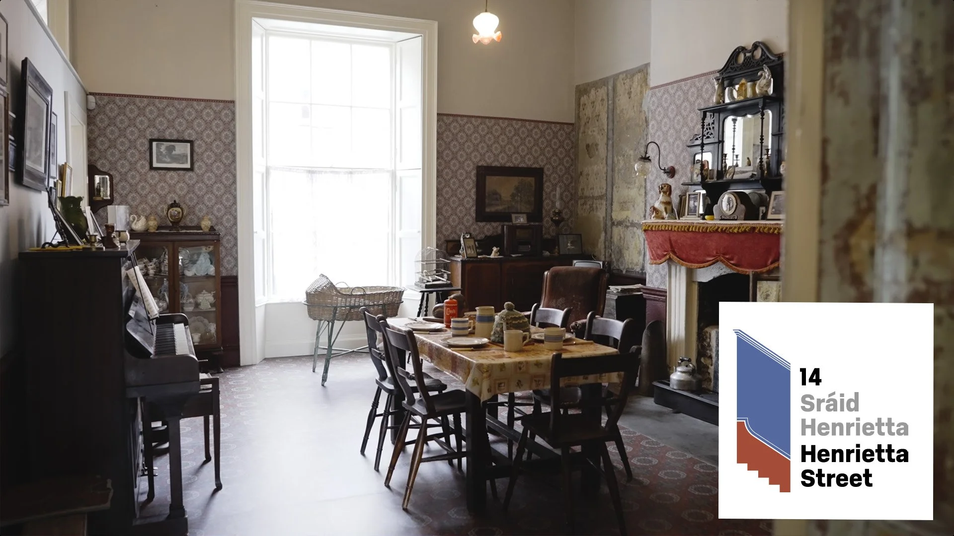 A cozy dining room with a large window letting in natural light. The room has vintage furniture, including a dining table with a tablecloth, several chairs, a piano, and various decorations on the walls and fireplace mantel. The walls are partly cove
