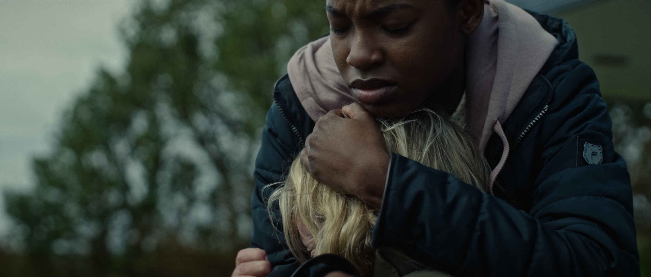 A woman with dark skin wearing a dark jacket and hoodie is holding a blonde-haired girl who is crying or distressed outdoors. The woman appears to be comforting her.