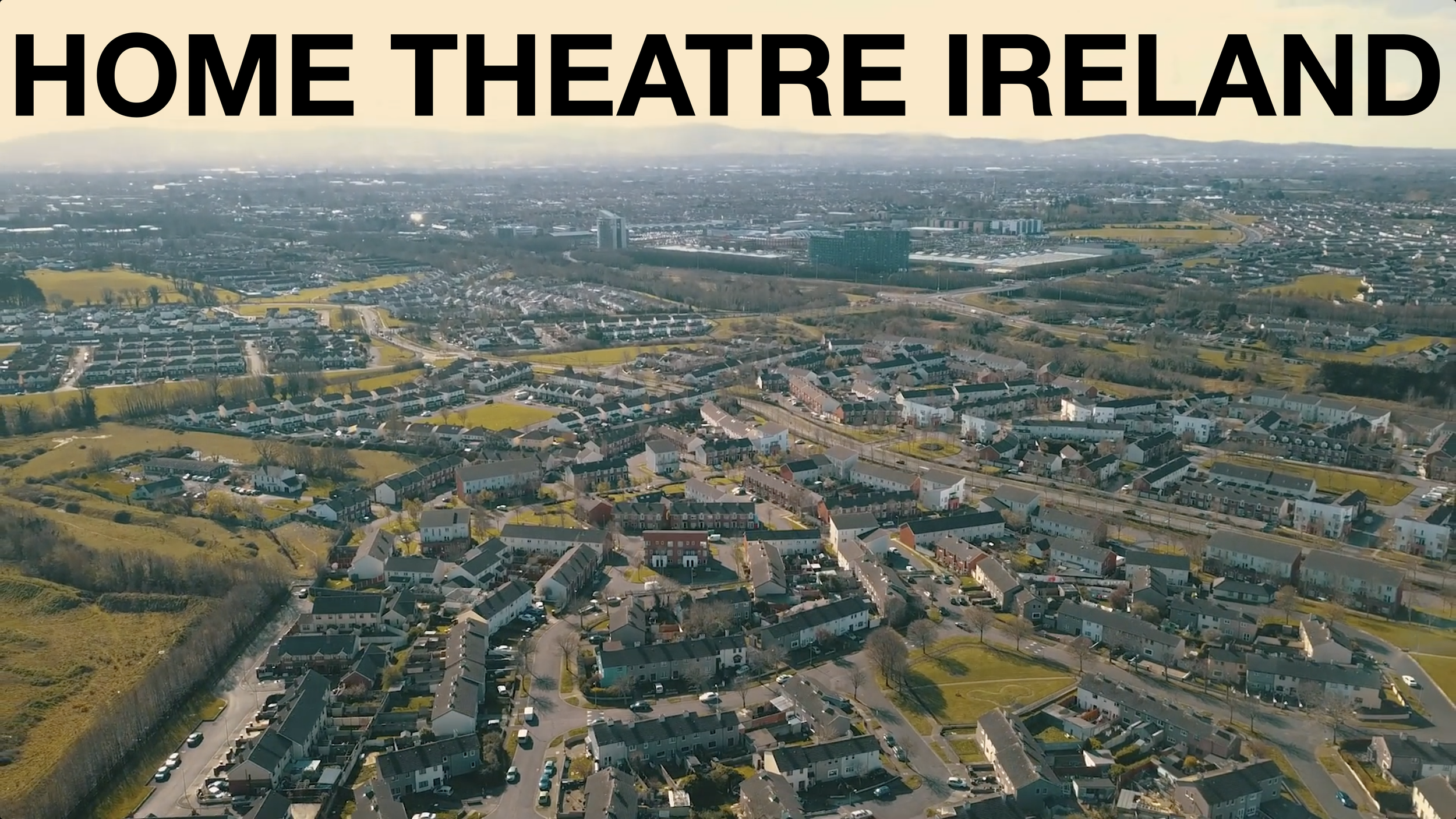 Aerial view of a suburban area in Ireland with houses, streets, and green fields, under a clear sky, with the text 'Home Theatre Ireland' overlaid at the top.