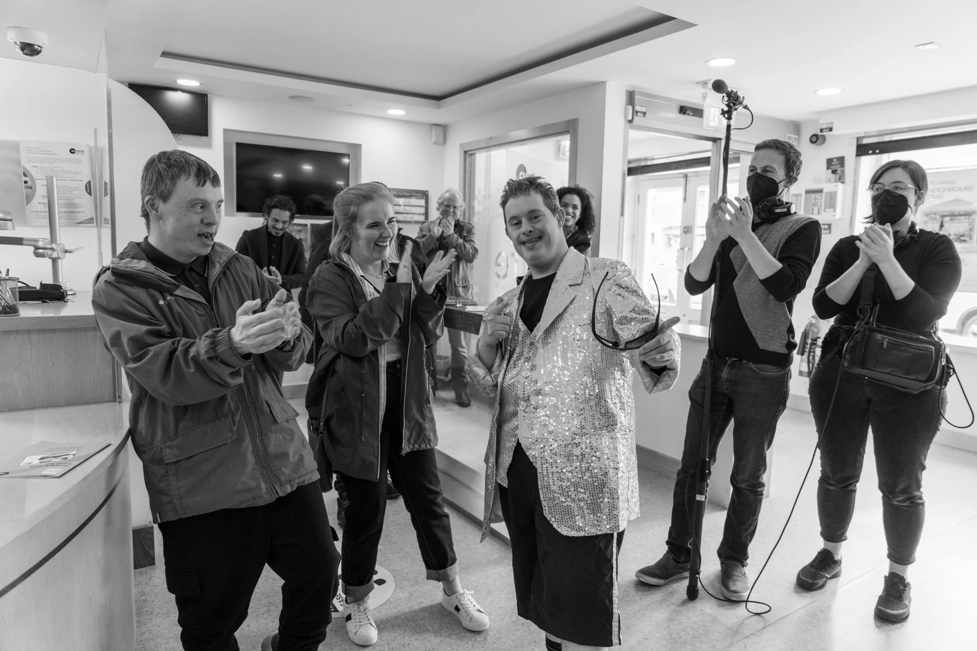 Group of people in a store clapping and smiling while a person dressed in a sparkly jacket holds sunglasses and poses.