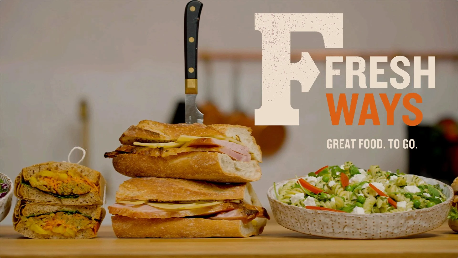 Assorted sandwiches, including a cut sandwich revealing a filling with vegetables, on a wooden surface. A bowl of pasta salad with vegetables is on the right. Overlay text reads 'F Fresh Ways' with 'Great food. To go.' underneath.