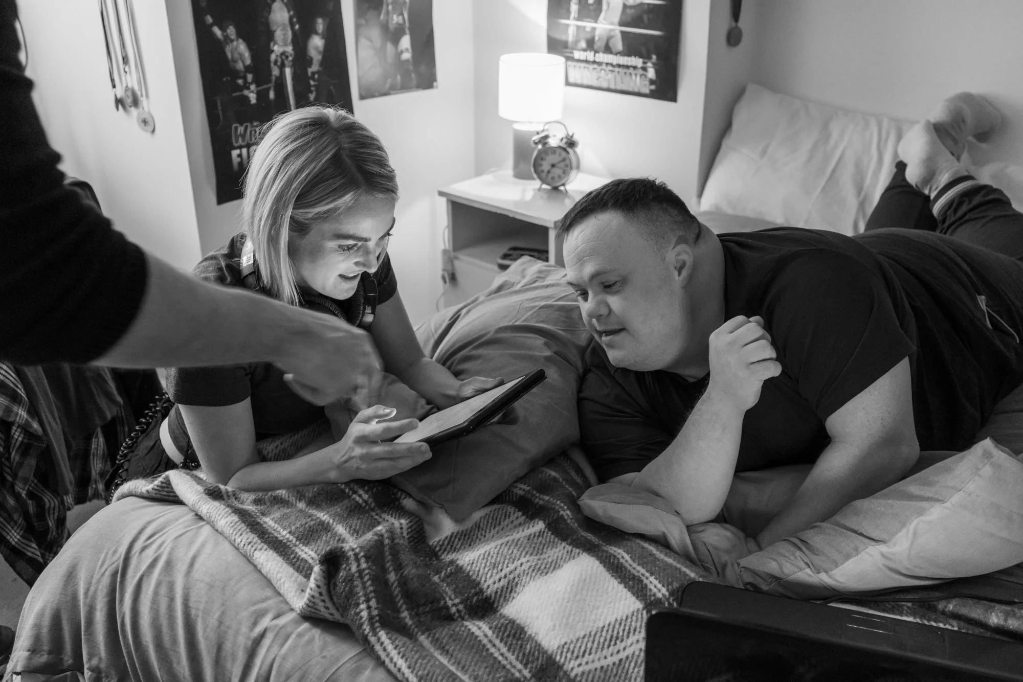 Behind the scenes photo. A woman and a man are lying on a bed looking at a tablet, with the woman smiling. An alarm clock and a lamp are on the nightstand beside the bed, and posters are on the wall in the background.