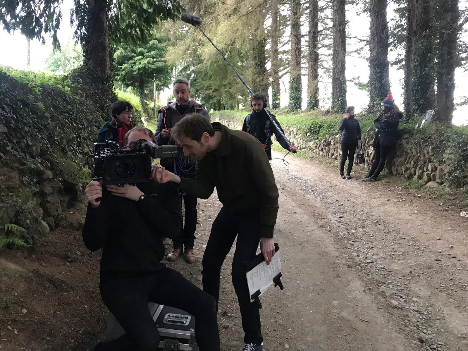 Filmmaking crew filming outdoors in a forested area with tall trees, some crew members are working with a camera, while others are setting up equipment or discussing.