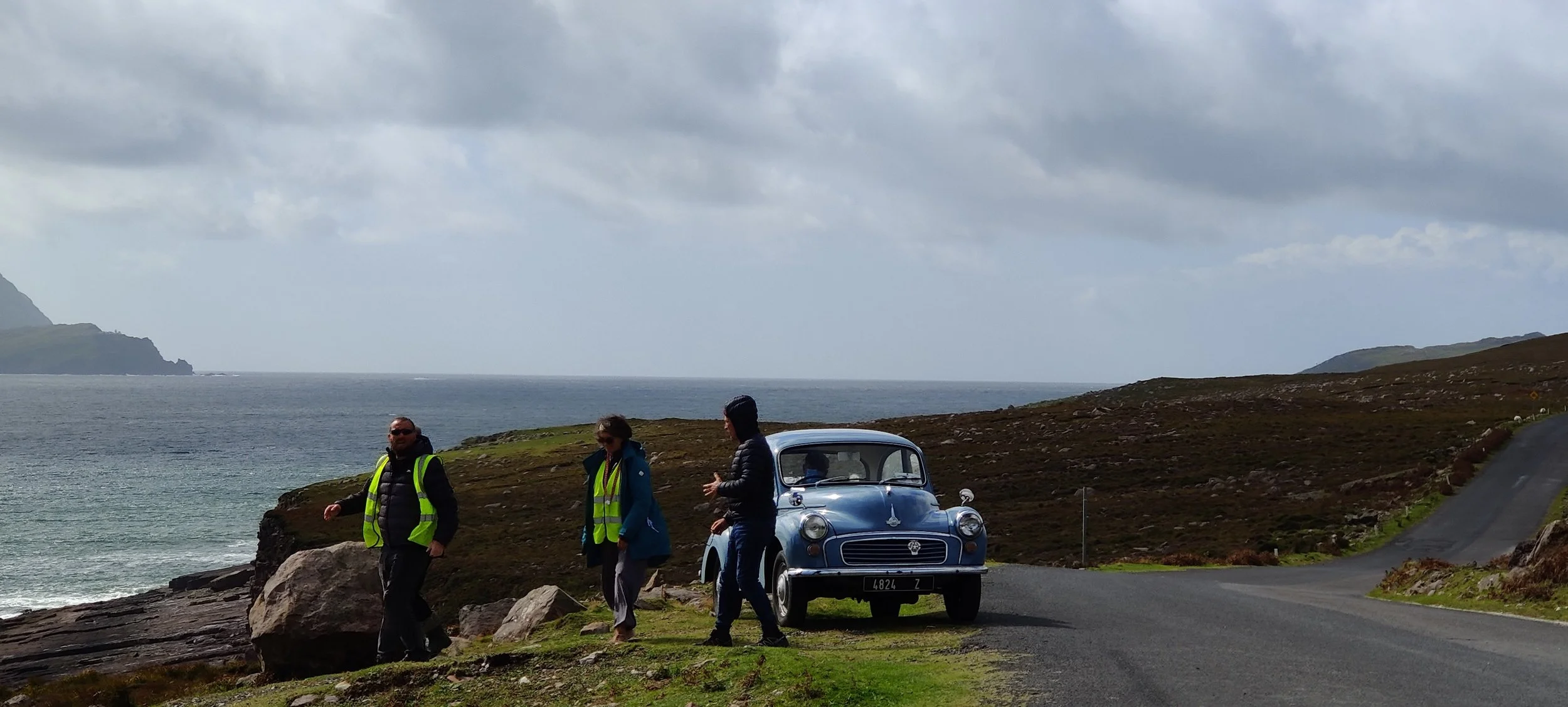 Three people standing near a vintage blue car on a coastal road with grassy landscape and ocean in the background, under cloudy sky.