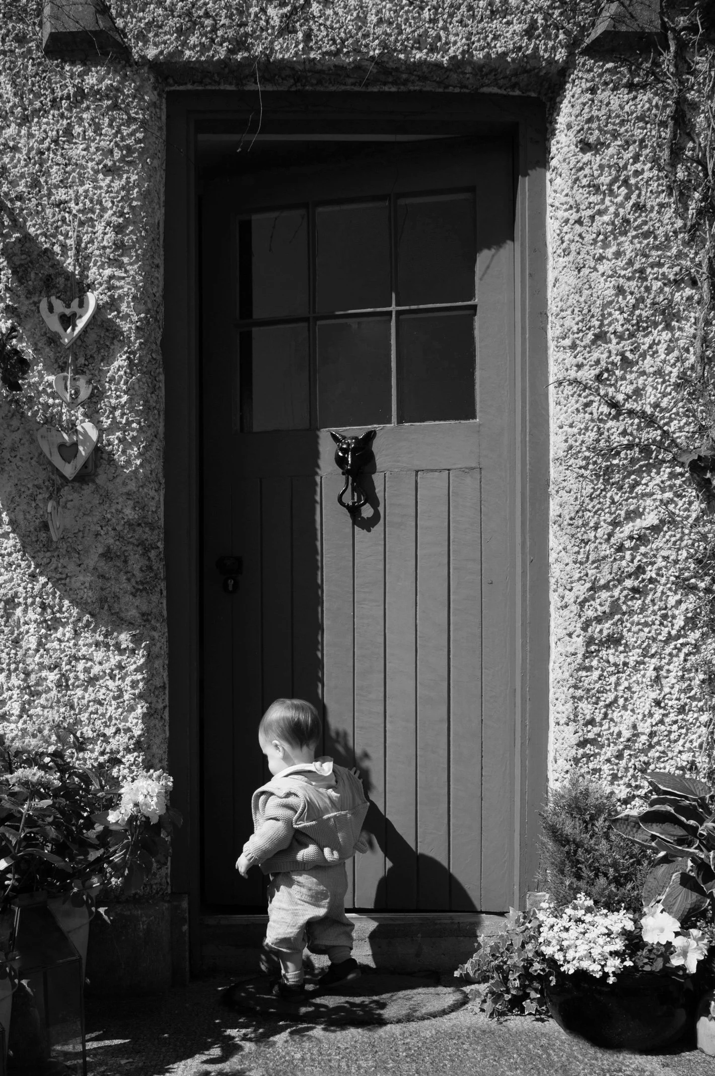 A young child wearing a hooded coat and boots is stepping out of a house onto a porch, in front of a wooden door with glass panes. The house has a textured exterior wall decorated with hanging heart-shaped ornaments and flowerpots on either side of t