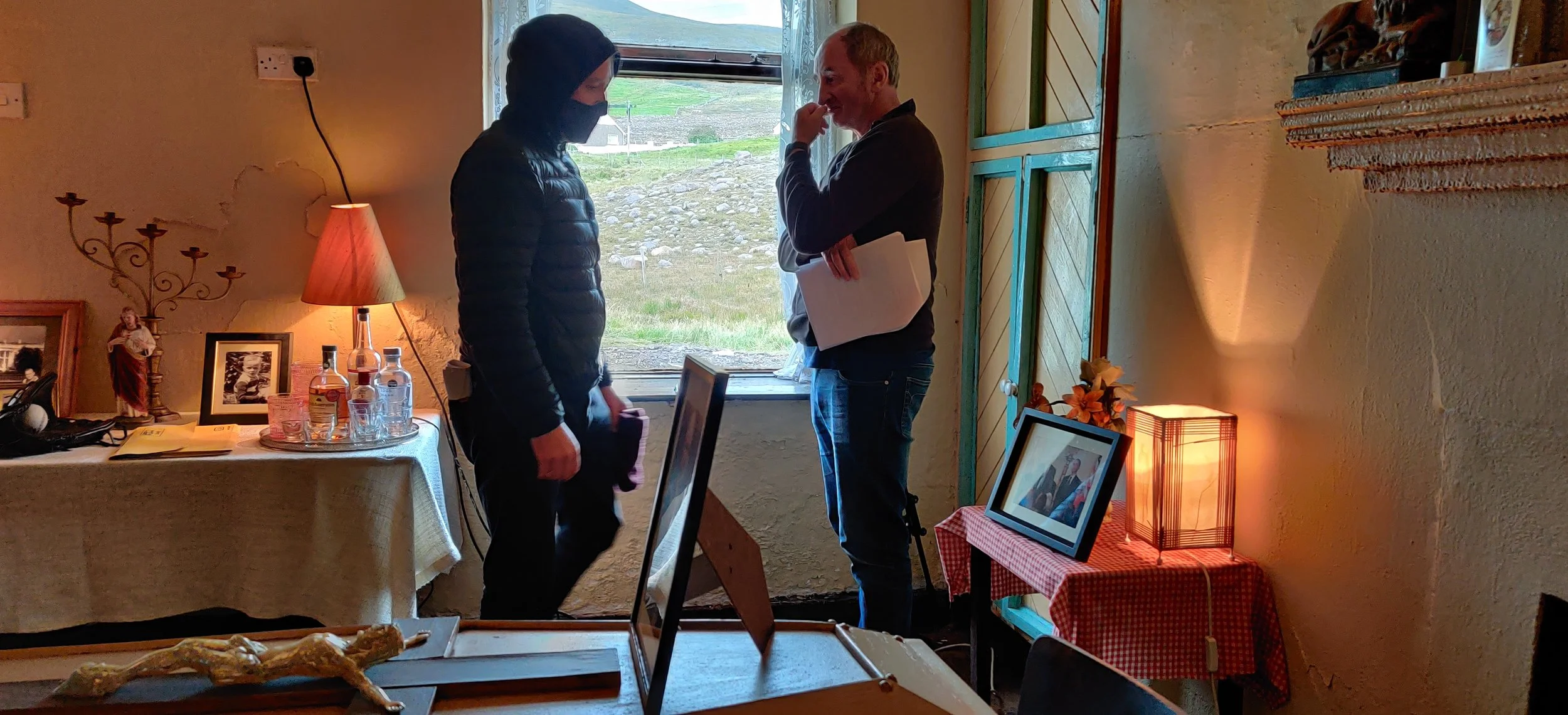 Two people standing near a window in a cozy room, engaged in a conversation. One person wears a mask and a black jacket, while the other holds a folder and is smiling. The room has warm lighting, framed photos, decorative items, and a table with bott