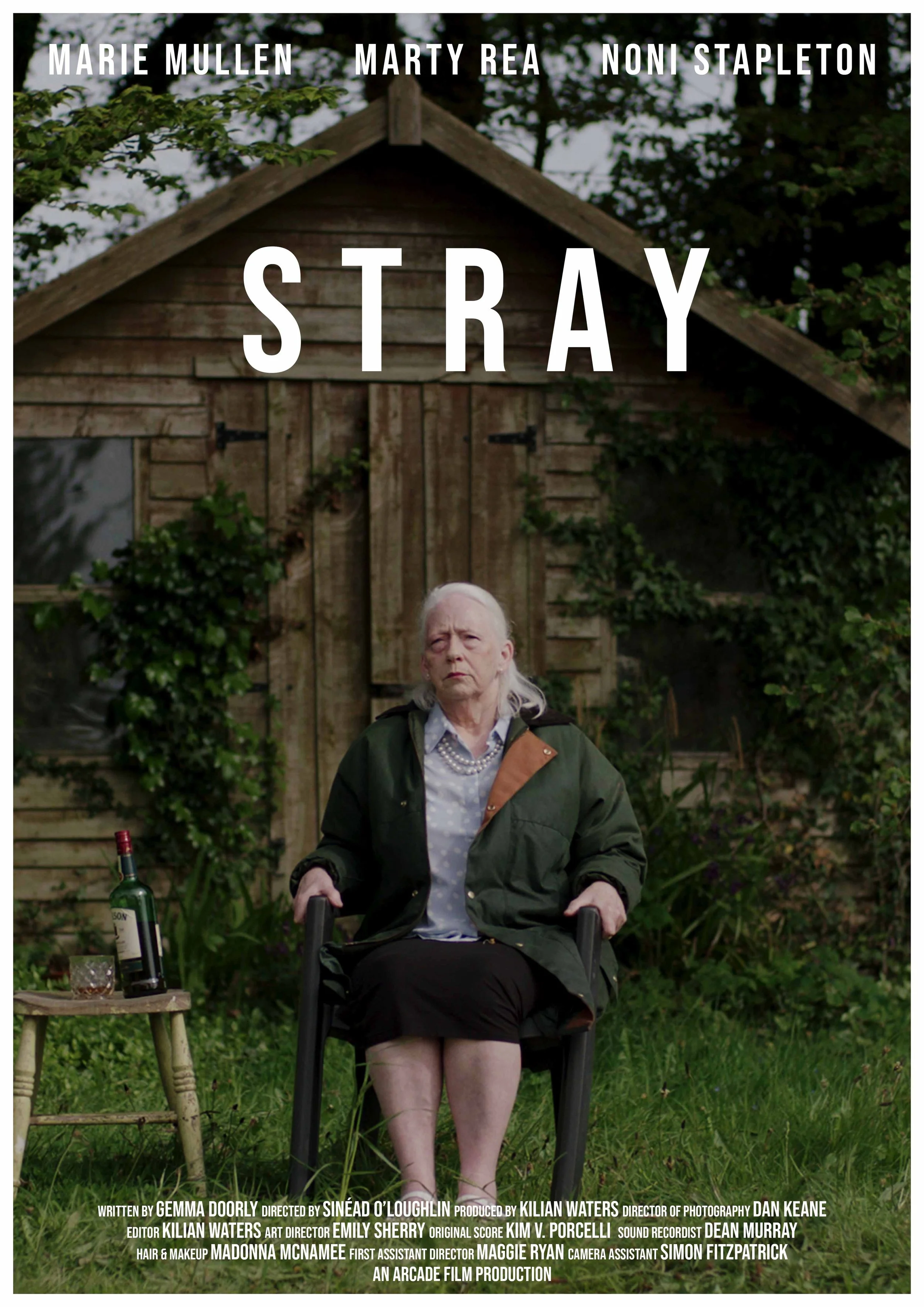 A woman sitting in a garden with a wooden shed in the background. She is holding onto the arms of a chair, has a serious expression, and appears older with white hair. There is a small wooden table beside her with a bottle of wine, a glass with wine,