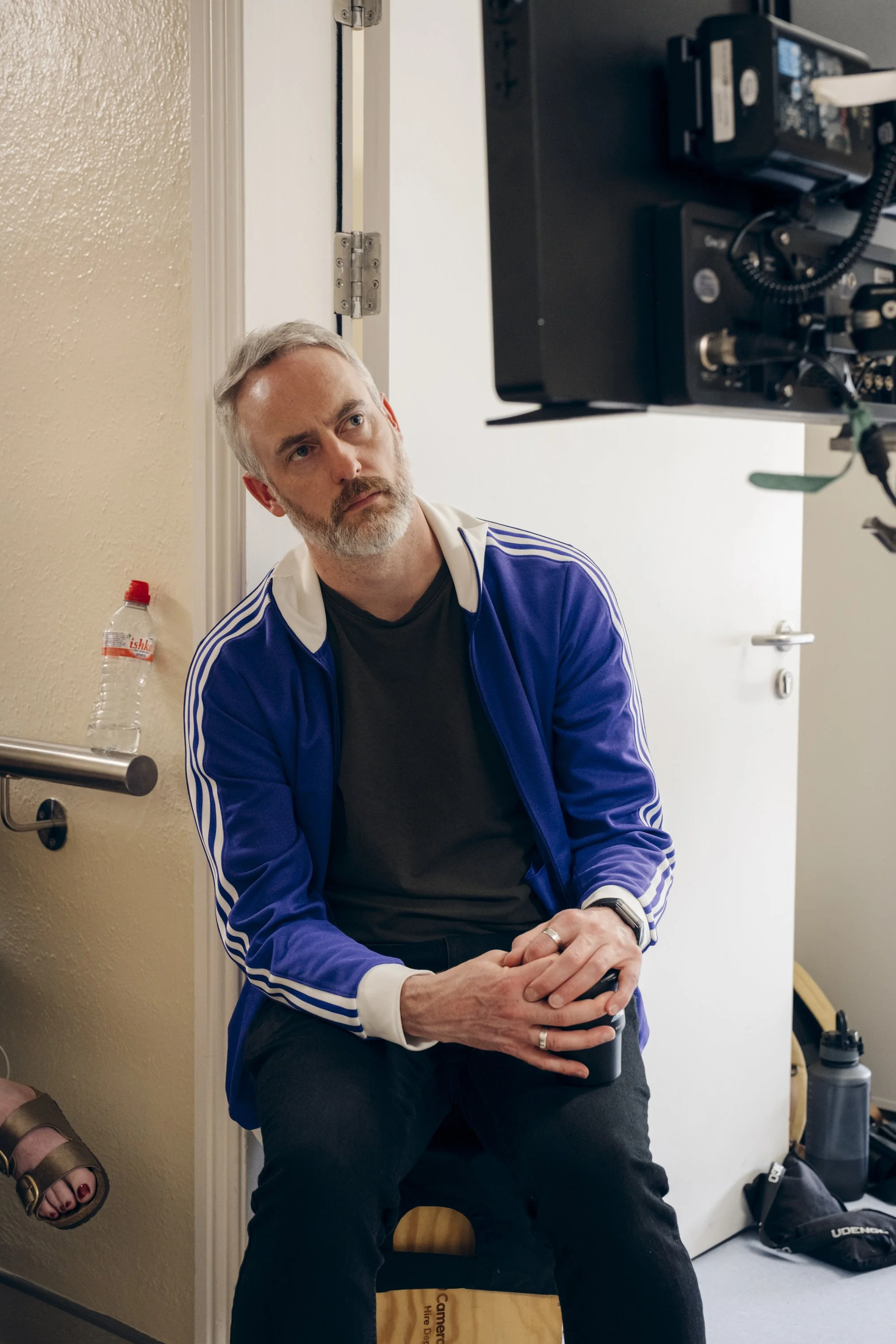 A middle-aged man with gray hair and a beard, wearing a blue jacket with white stripes, seated and looking to the side. Behind him, there is a white wall, a partially open door, and a black camera or recording device mounted on the wall. A plastic wa