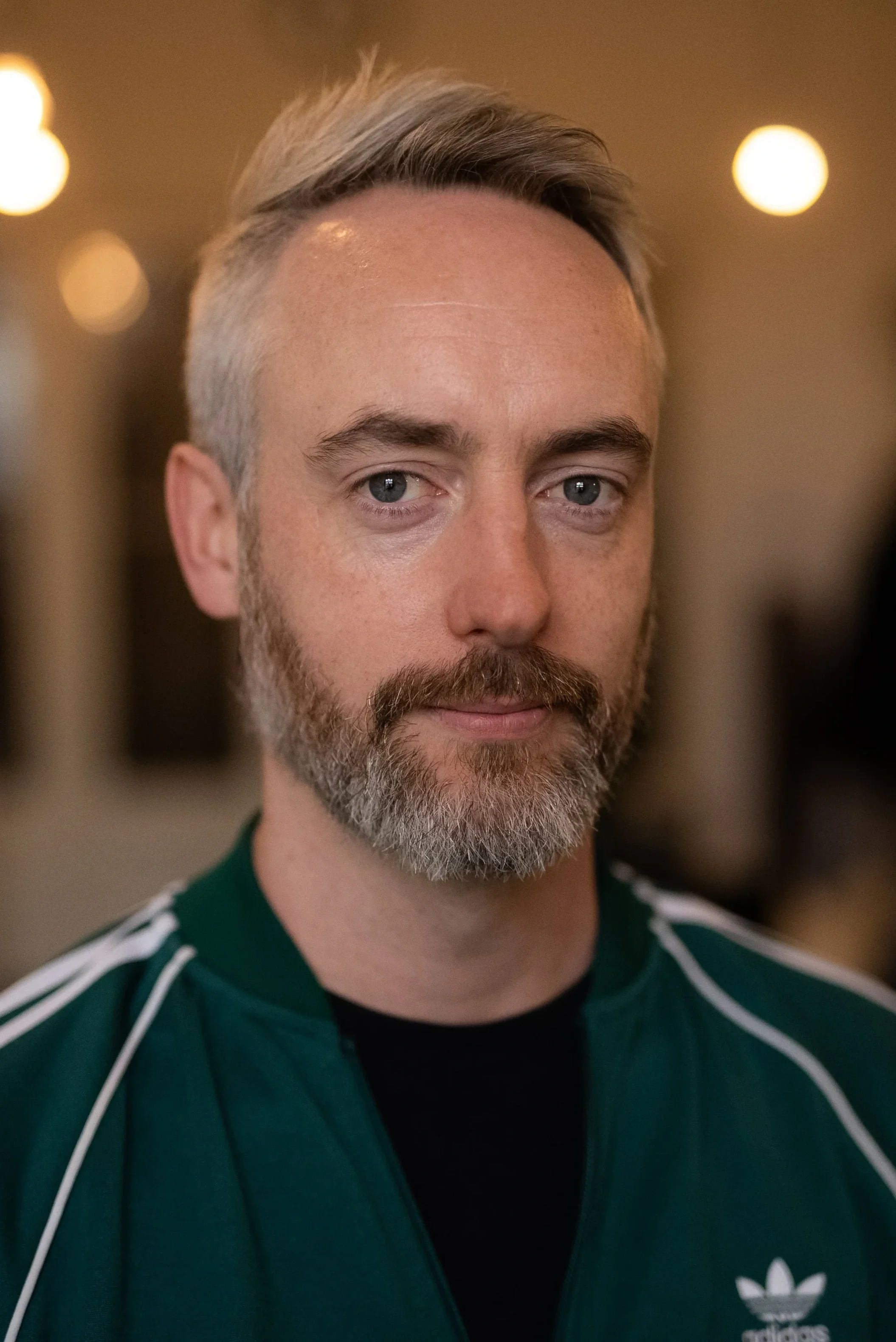 Close-up of a man with short gray hair and beard, wearing a dark green Adidas jacket with white stripes, looking at the camera indoors with blurred warm lights in the background.