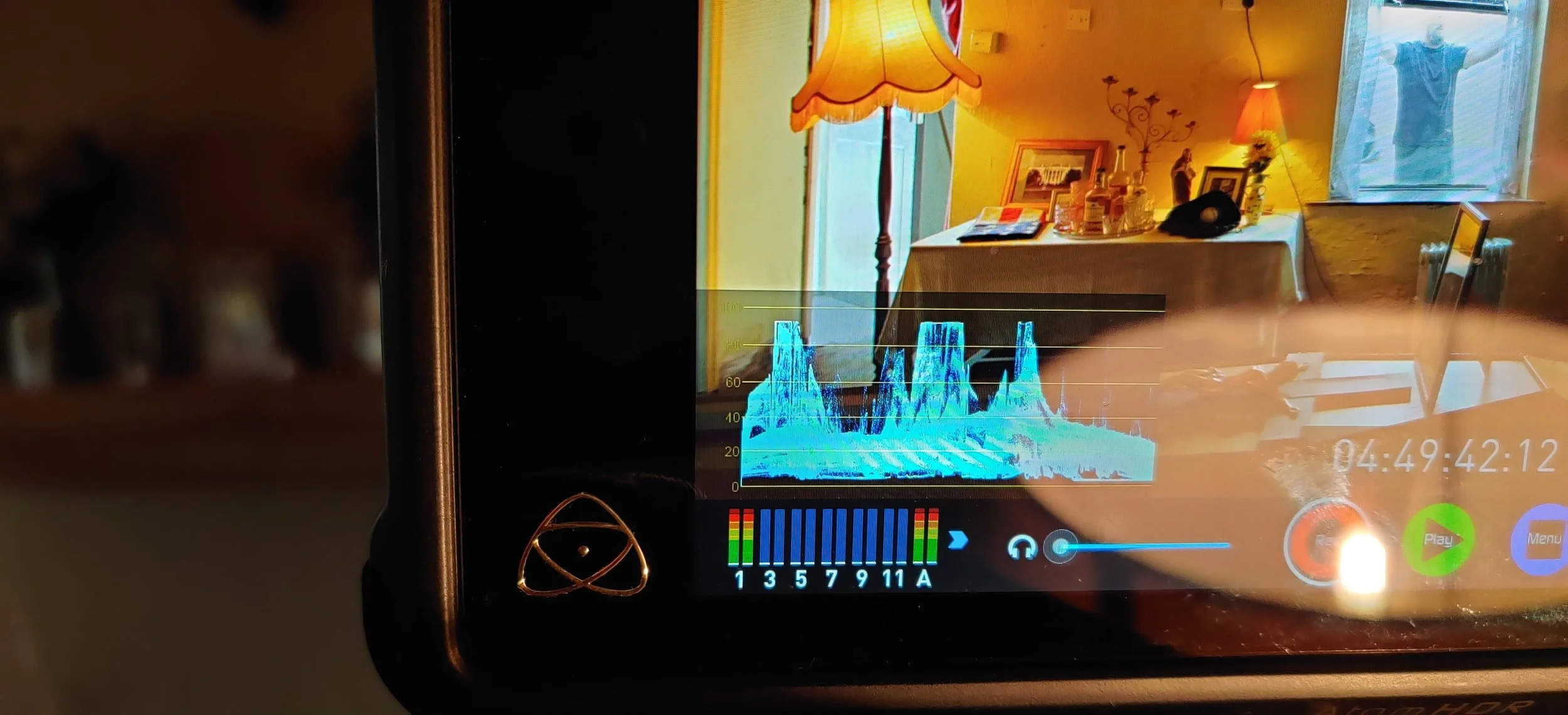 Behind the scenes photo. A digital screen displays a color-coded graph, with a background of a cozy room having yellow walls, a wooden table with decorative bottles, framed photos, a table lamp, and window curtains.