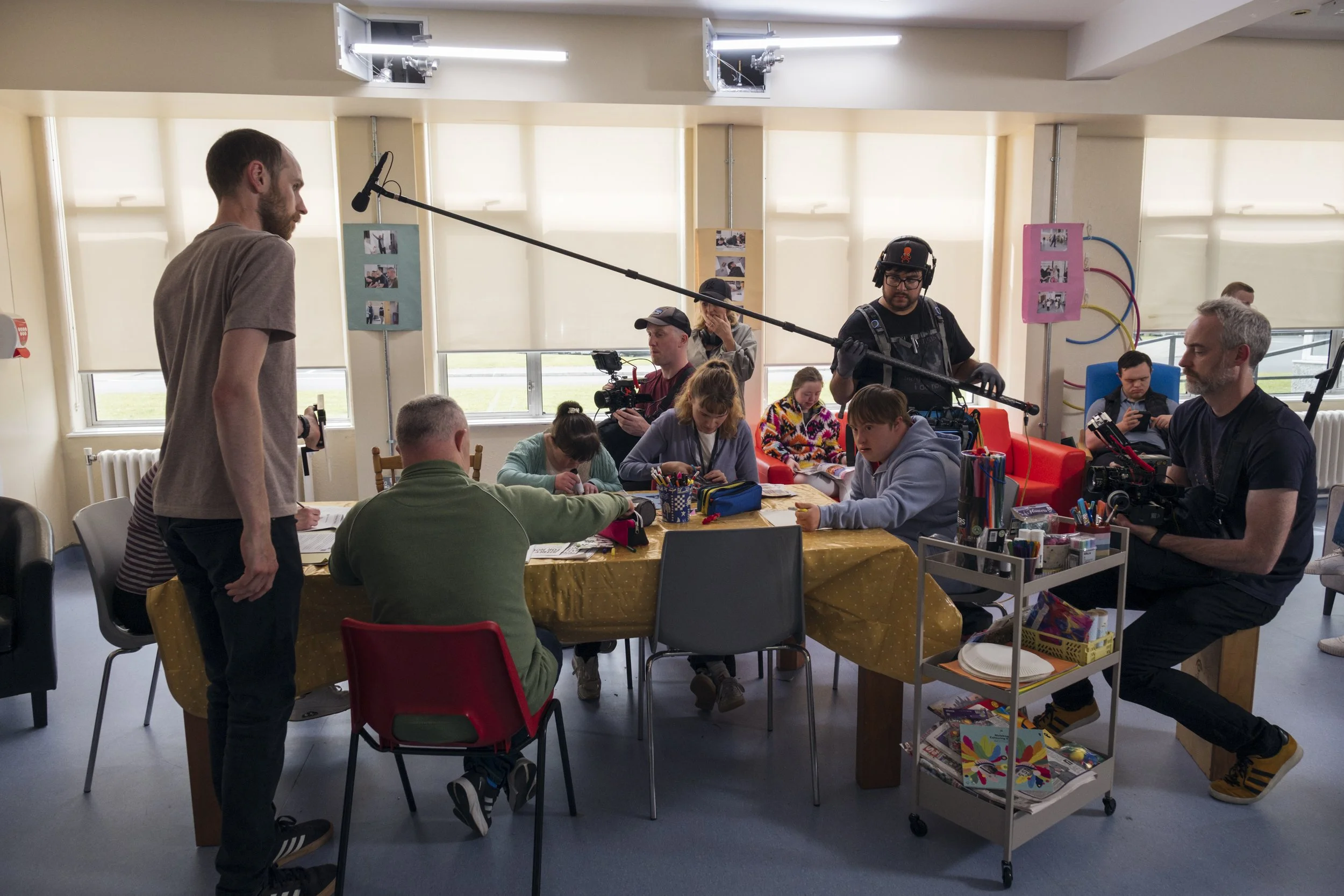 Filmmaking crew recording a scene at an indoor location with a boom microphone, camera operators, and various crew members seated around a table filled with supplies.