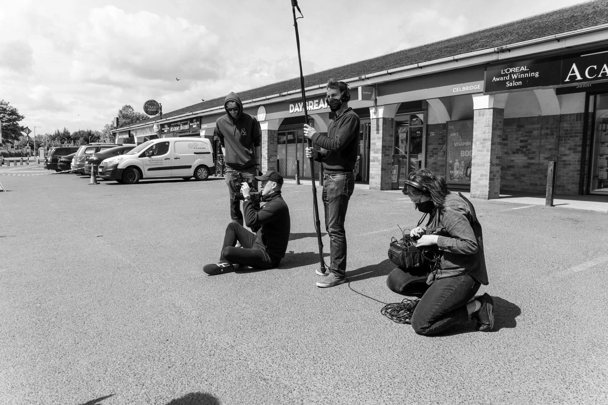 A film crew shooting a scene outdoors in a parking lot. One person is sitting cross-legged with a camera, while another is kneeling handling equipment. Two men stand, one holding a boom microphone and another with headphones and a face mask. Shops an