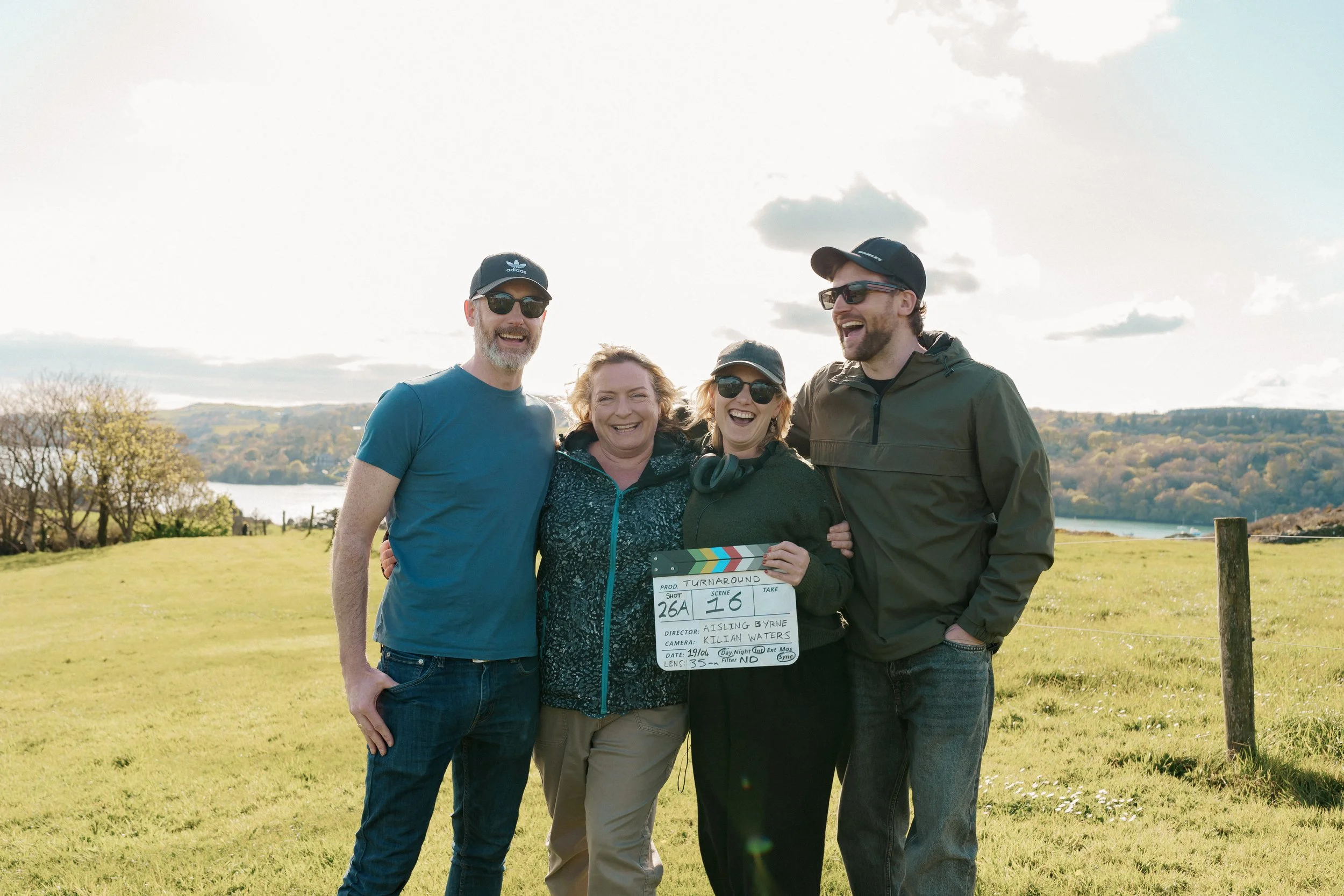 Four people standing outdoors on a sunny day, smiling and posing for a photo, holding a clapperboard, with trees, a body of water, and a sky with clouds in the background.