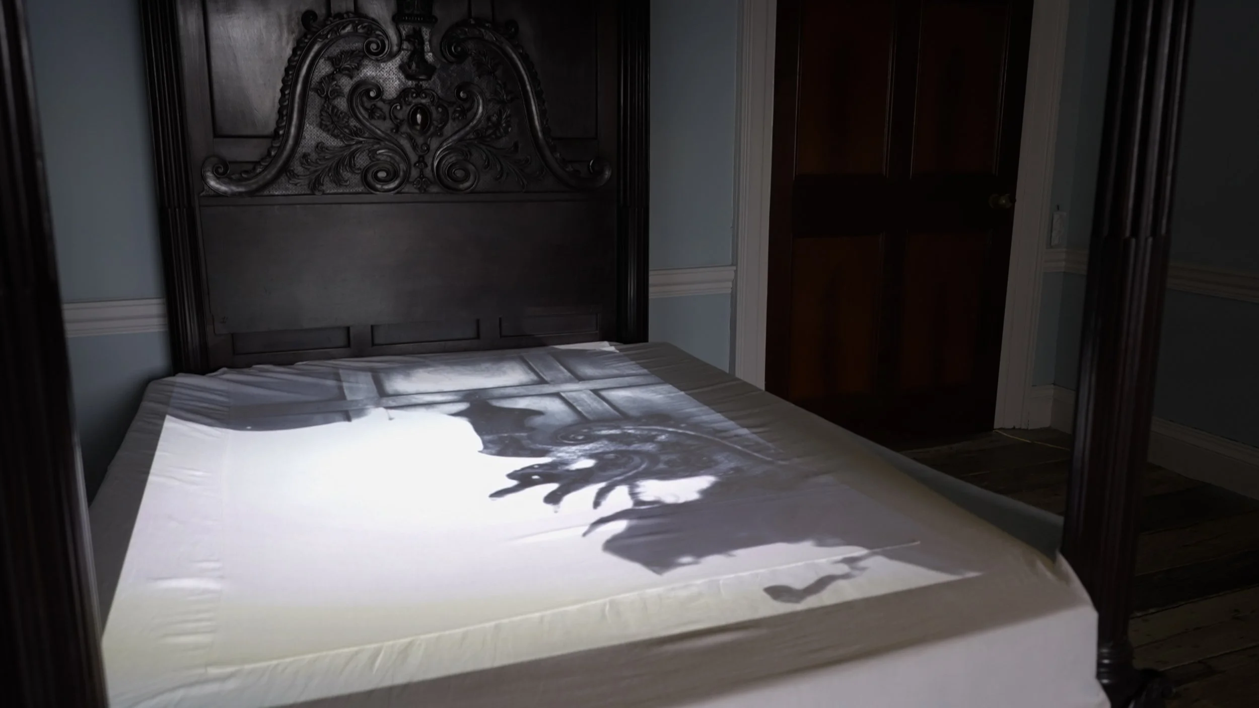 A dark wooden bed with an ornate carved headboard, a white bed sheet, and shadows of leaves cast on the sheet in a dimly lit room.