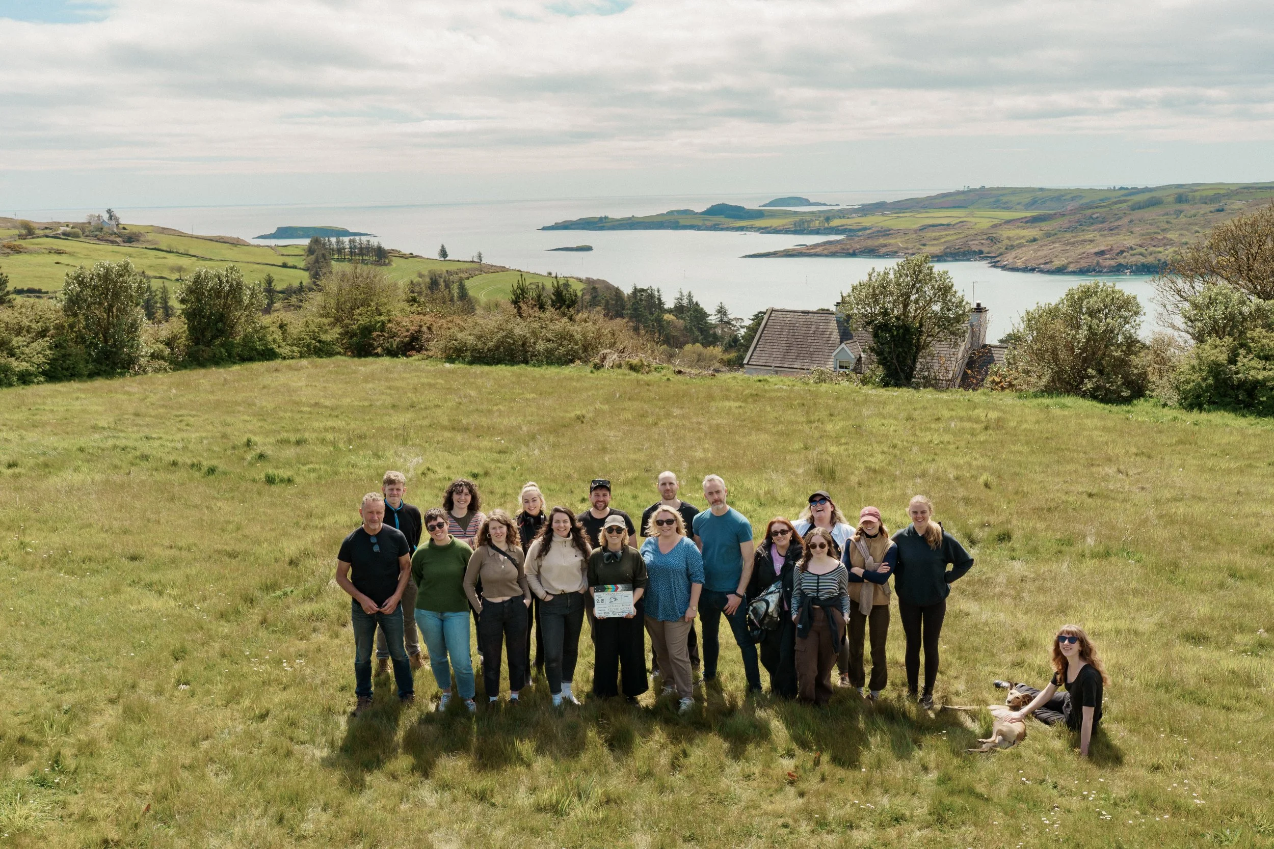 Group of people standing in a grassy field overlooking a lake and hills, with cloudy sky.