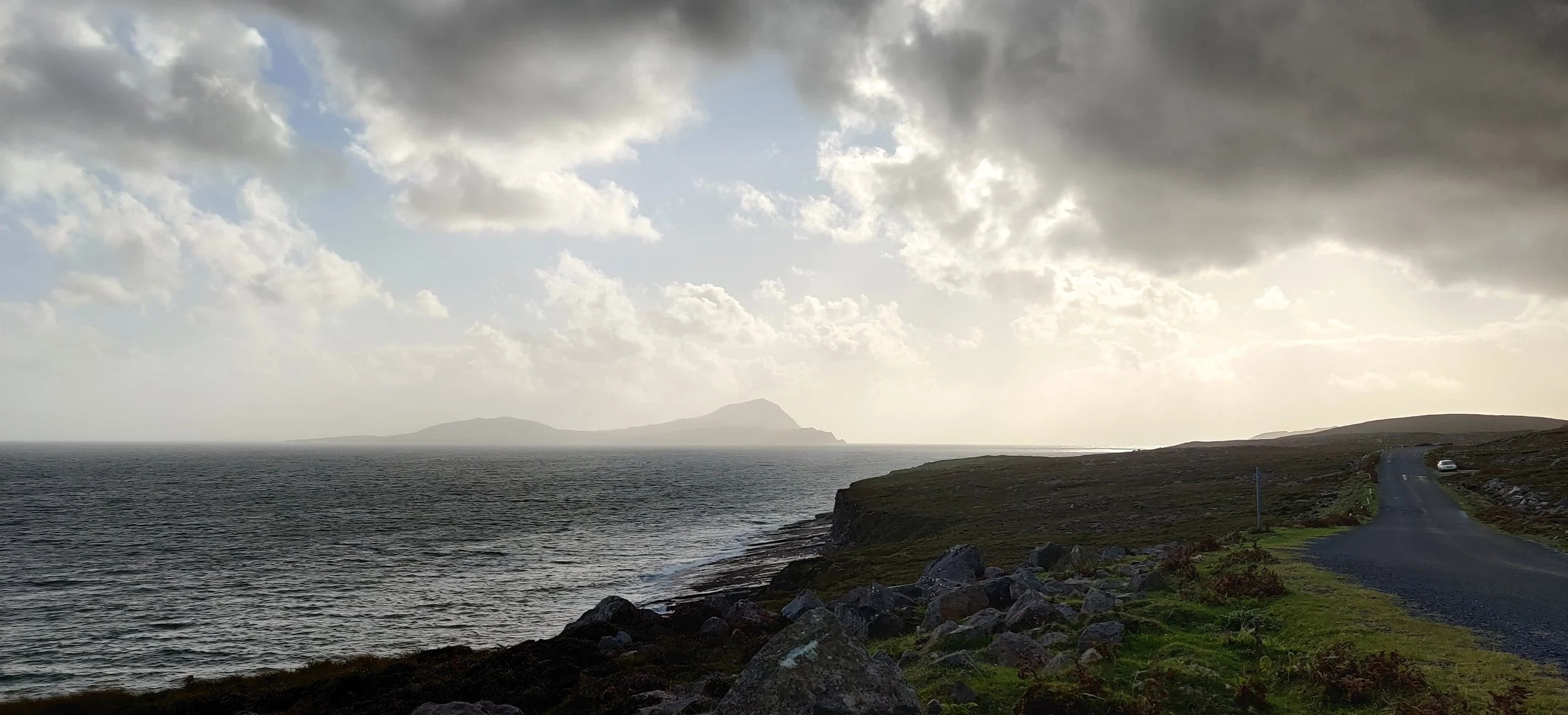 Coastal landscape with a winding road along the rocky shoreline, ocean waves, distant island, and a cloudy sky with patches of sunlight.