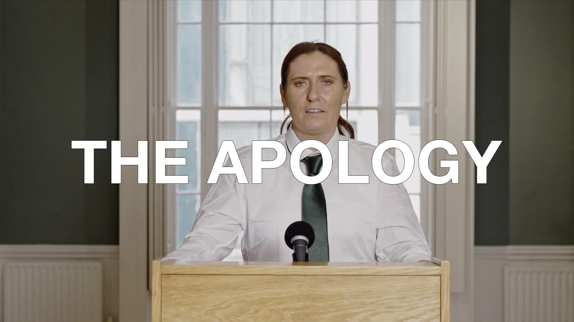 A woman with brown hair tied back, wearing a white shirt and a dark tie, stands behind a wooden podium with a microphone. She appears to be giving a speech or statement, with a large window in the background. The words 'THE APOLOGY' are superimposed 