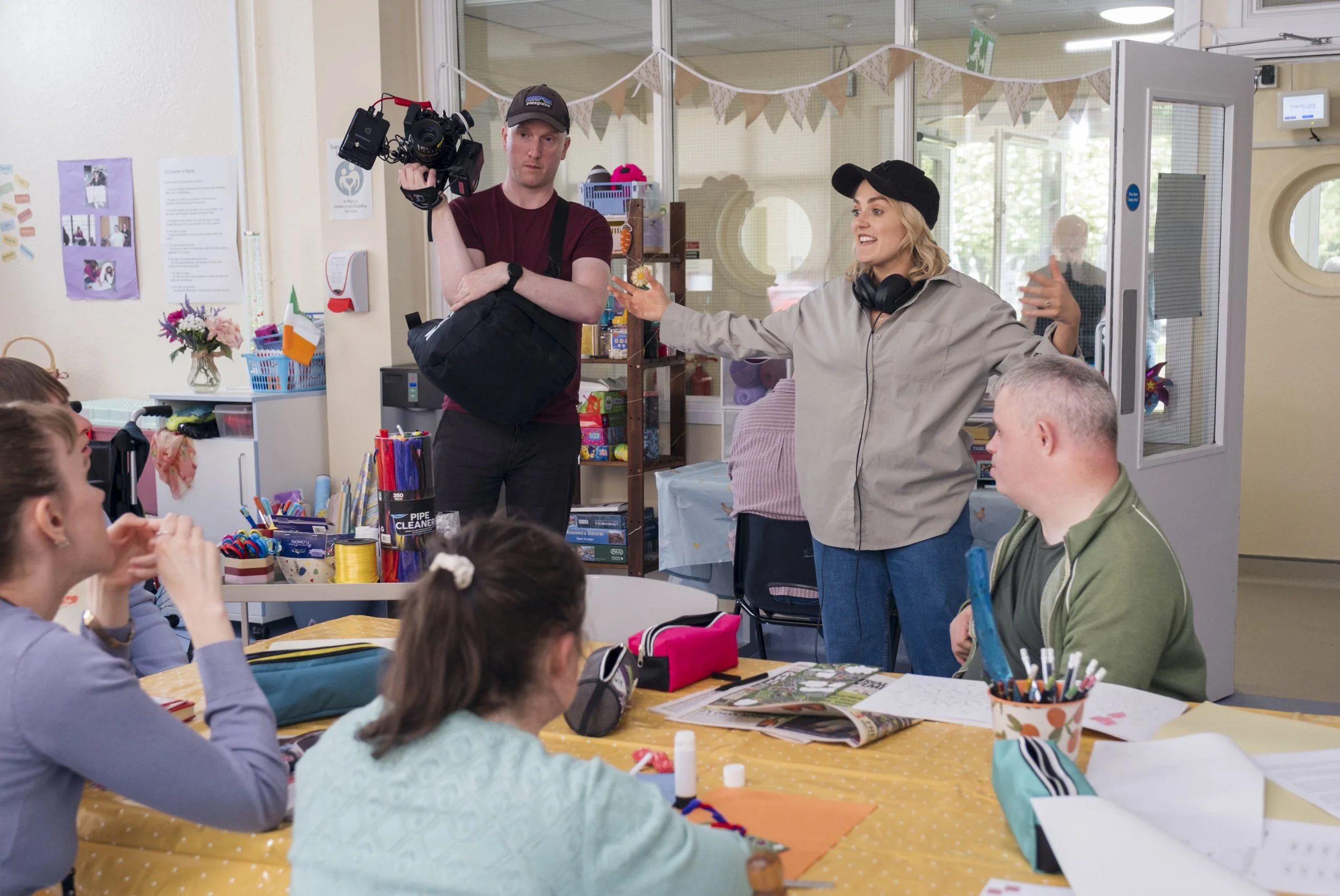 A woman in a gray jacket and black cap stands at a table with children, talking and gesturing with her hands, while a man in a maroon shirt, black cap, and carrying a camera on his shoulder watches her. The setting appears to be a classroom or craft 