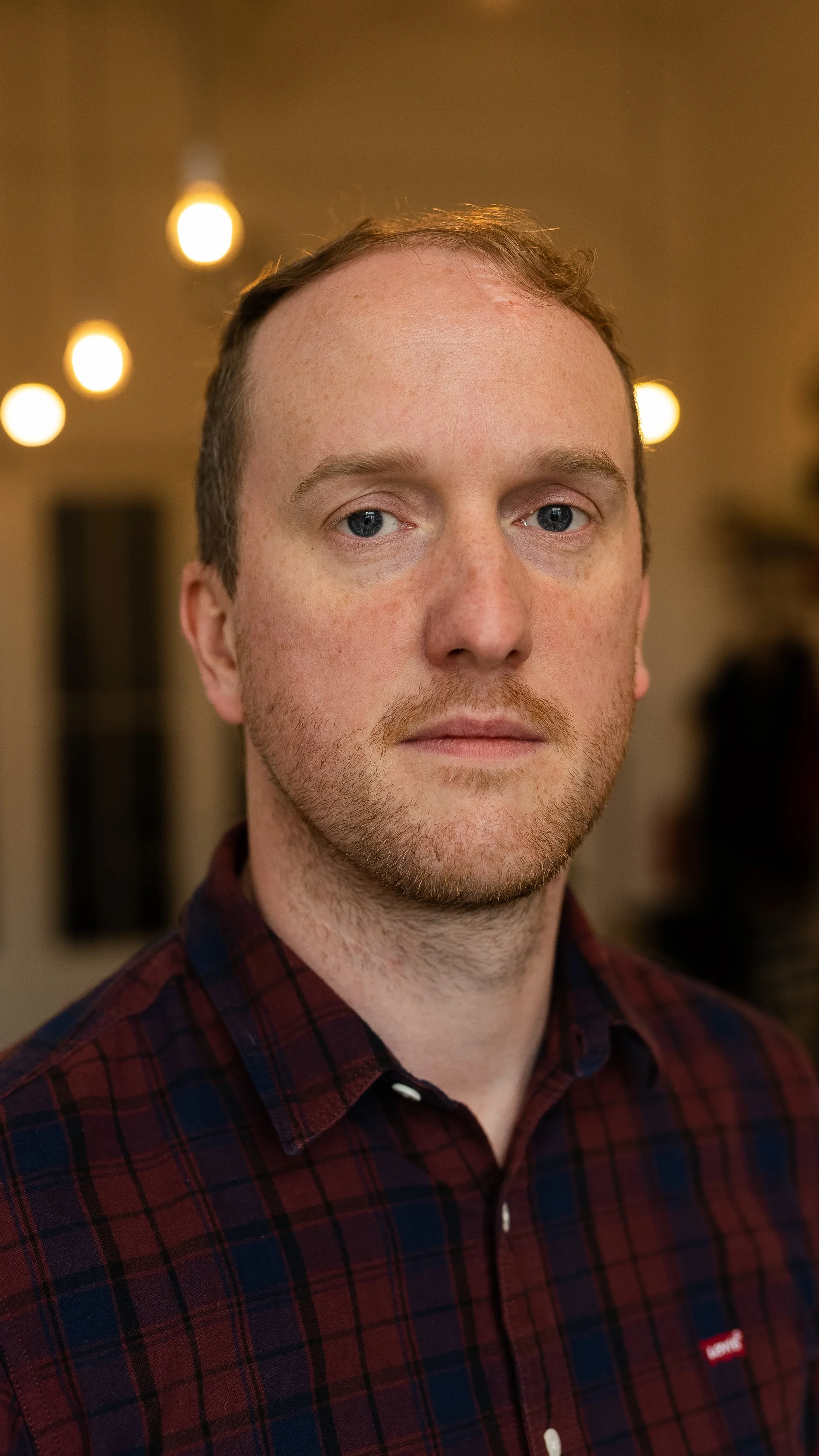 A portrait of a man with reddish hair and beard wearing a red and blue plaid shirt, standing indoors with blurred warm lighting in the background.