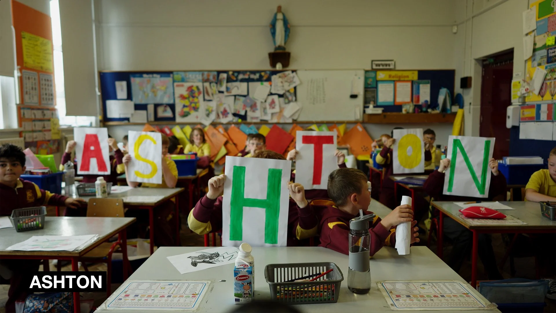 Classroom of young students holding up large letters that spell 'ASHON' during a classroom activity.
