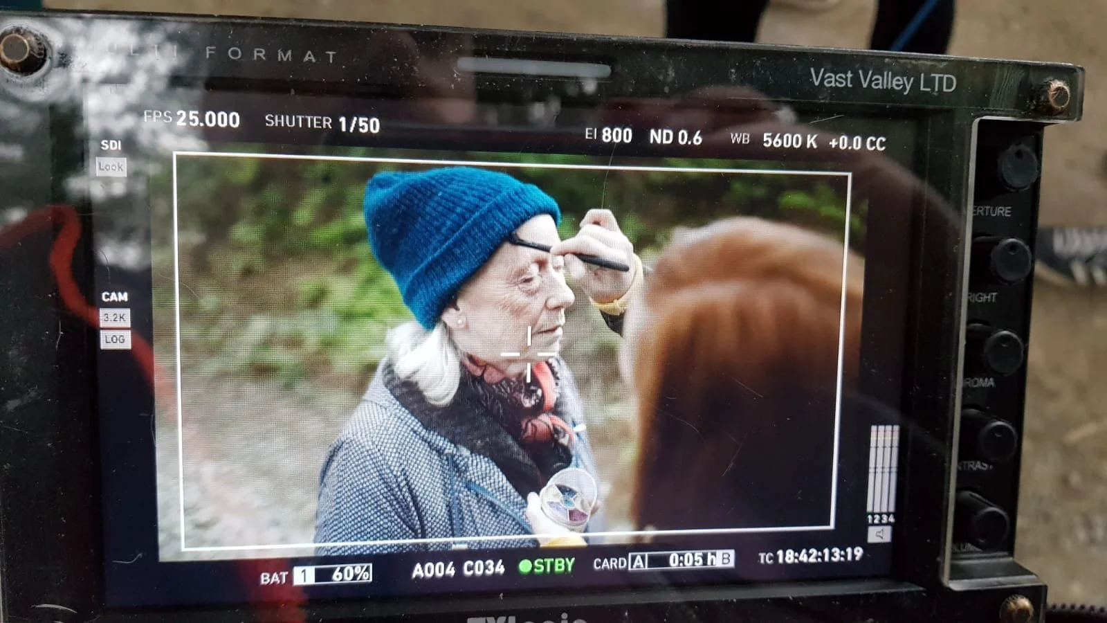 A behind the scenes photo. A woman with gray hair and a blue knit cap having makeup or face paint applied by another person outdoors.