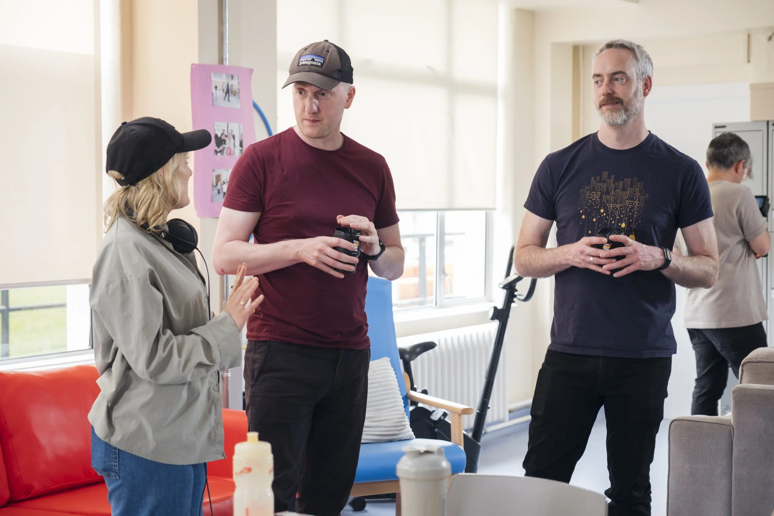 Three people are engaged in a conversation in a bright room with large windows. The woman on the left is wearing a black cap and headphones around her neck, speaking to two men. The man in the middle is wearing a burgundy shirt and a gray cap, holdin