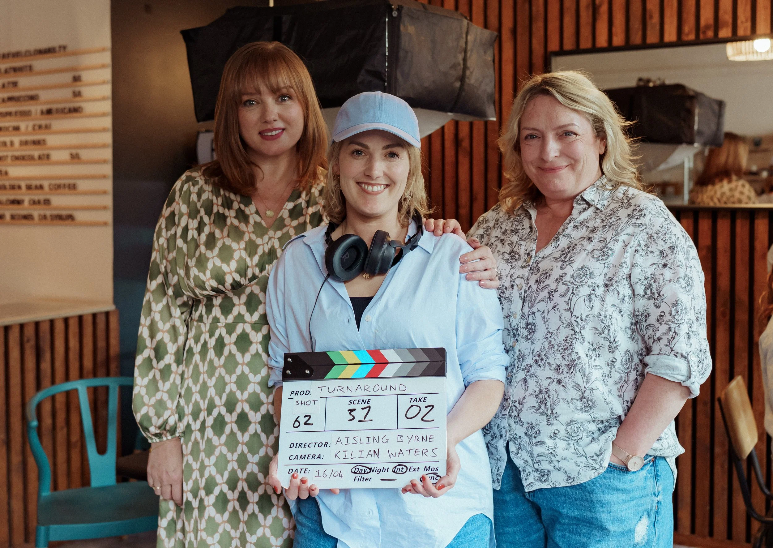 Three women standing indoors, one holding a film slate. The woman in the middle is wearing a light blue shirt, a blue baseball cap, and has headphones around her neck. The woman on the right has blonde hair and is wearing a floral shirt. The woman on
