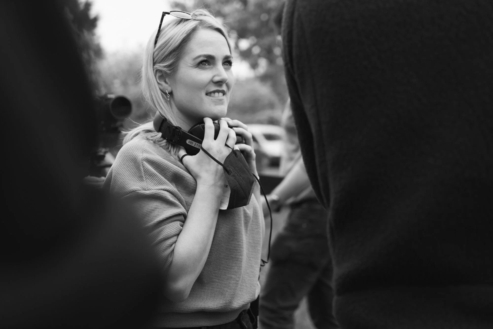 Behind the scenes photo from a film set. A smiling woman with blonde hair wears glasses on her head and a glad-sleeved top. She holds a walkie-talkie and appears to be engaged in conversation, surrounded by other people in an outdoor setting.
