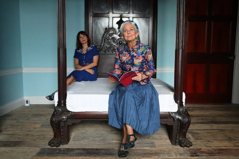 An elderly woman sitting on a four-poster bed holding a book, with a young woman sitting on the bed in the background. The room has blue walls, wooden flooring, and dark wooden furniture.