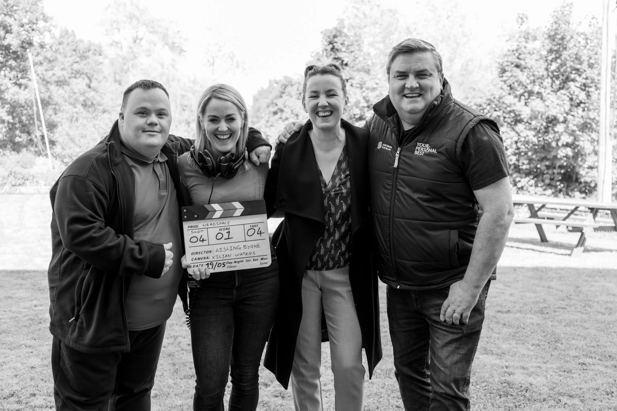 Behind the scenes photo. Five people smiling and standing outdoors on a grassy field. One person is holding a film slate. Four people have their arms around each other. The background has trees and a picnic table. The photo is in black and white.