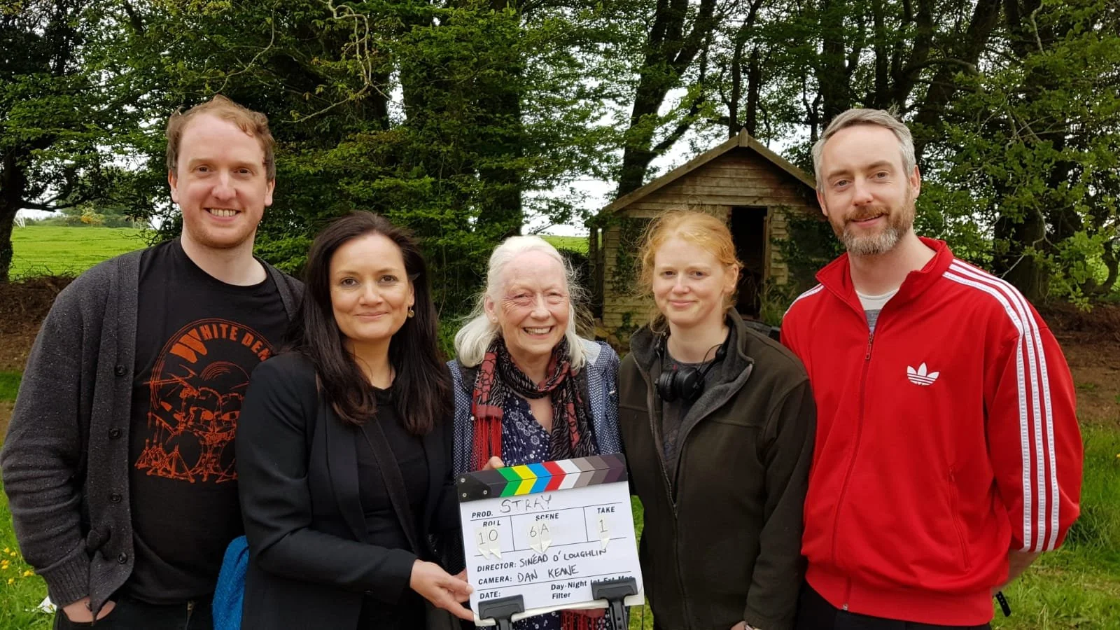 Group of five people standing outdoors on a film set, with greenery and a small wooden structure in the background, holding a clapperboard.