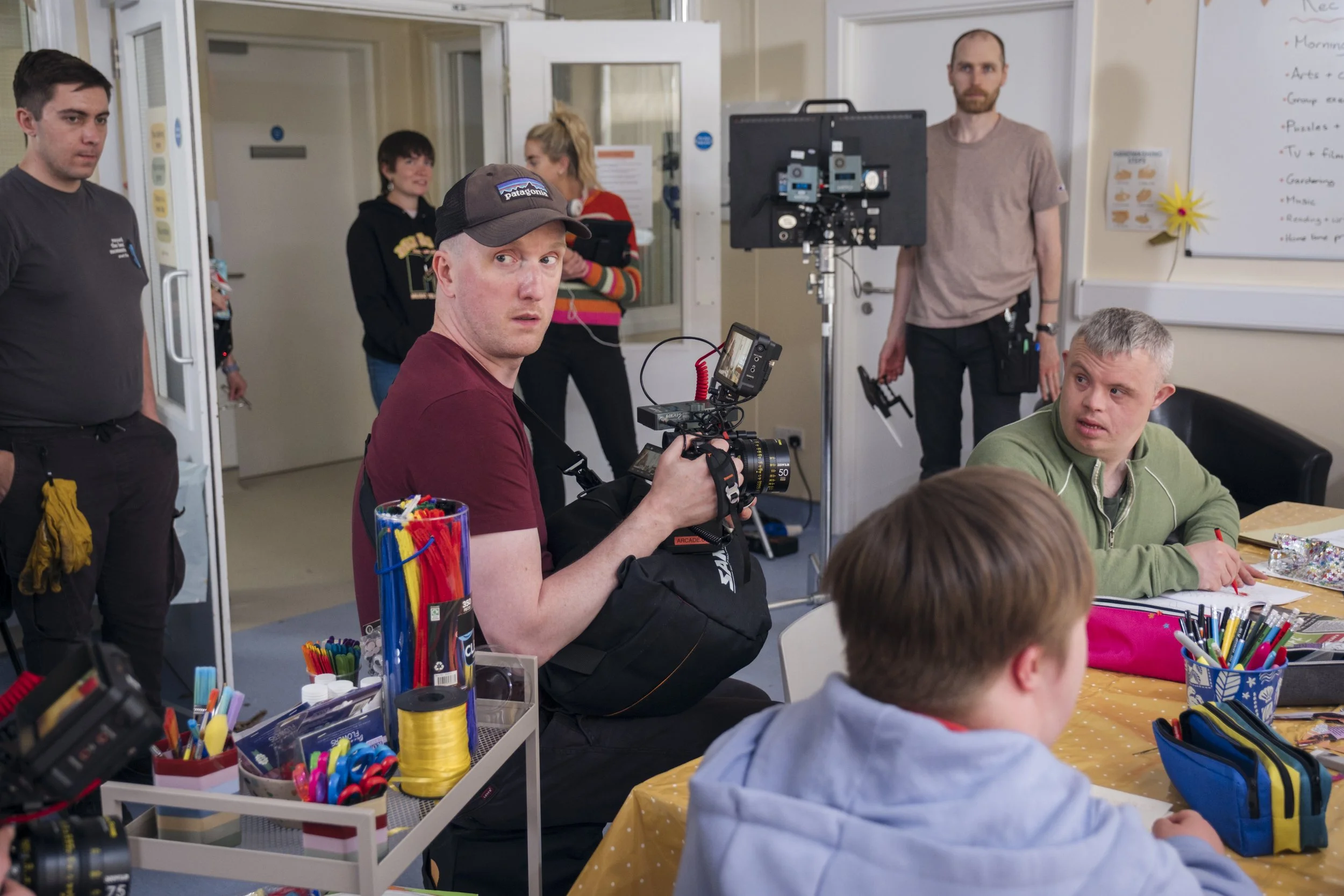 People working on a film set or video production inside a classroom or meeting room, with equipment like cameras, monitors, and colorful art supplies on the table.