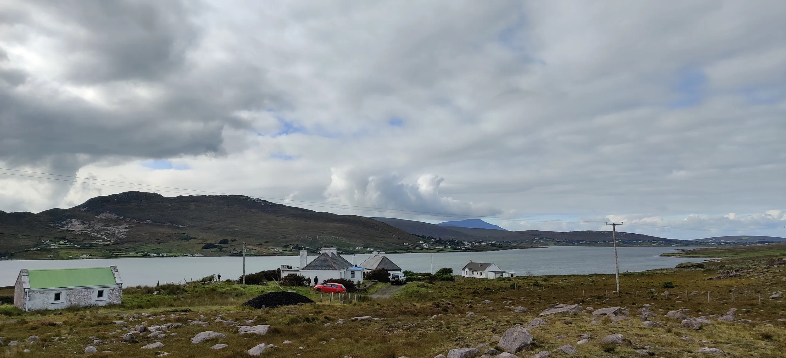 A rural landscape with a large body of water, hills, and houses, under a cloudy sky.