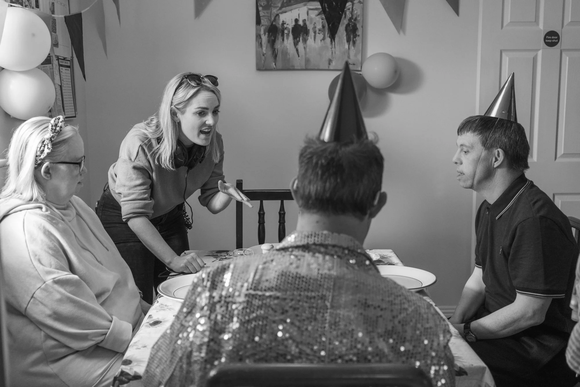 Behind the scenes photo from a film set. A woman speaking to three seated people at a table during a birthday celebration, with party hats and balloons as decorations.