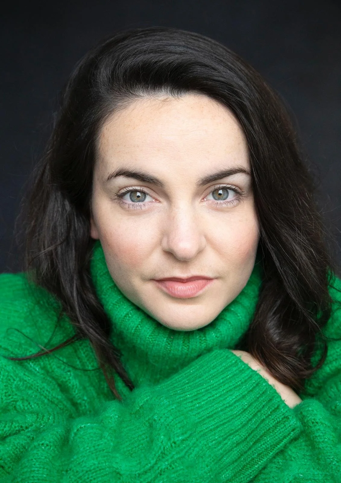Close-up portrait of a woman with blue eyes and dark brown hair wearing a bright green turtleneck sweater against a dark background.